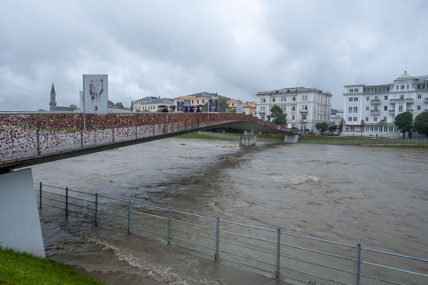Marko Feingold Steg pedestrian bridge spans the flooded Salzach river under grey skies in Salzburg.
