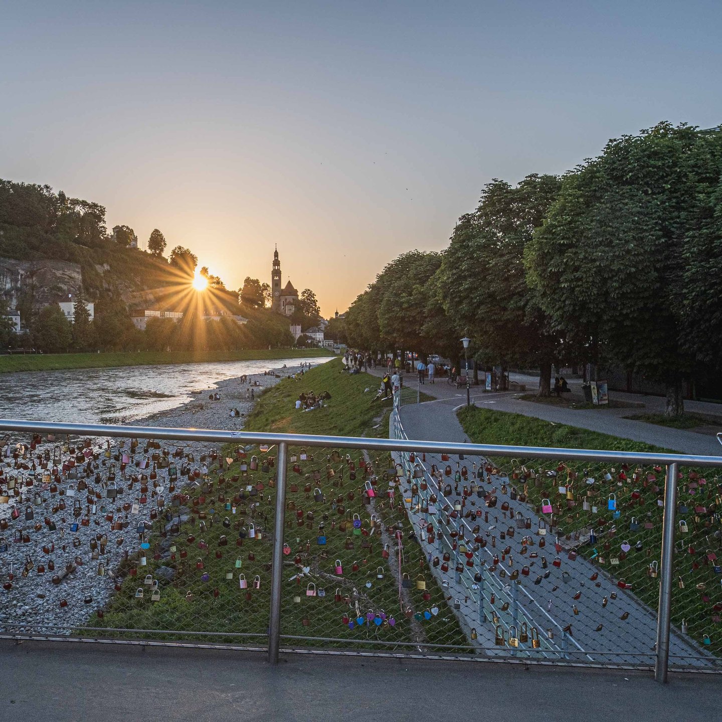 Love locks on Marko Feingold Steg railing with Salzach river and church tower at sunset in Salzburg.