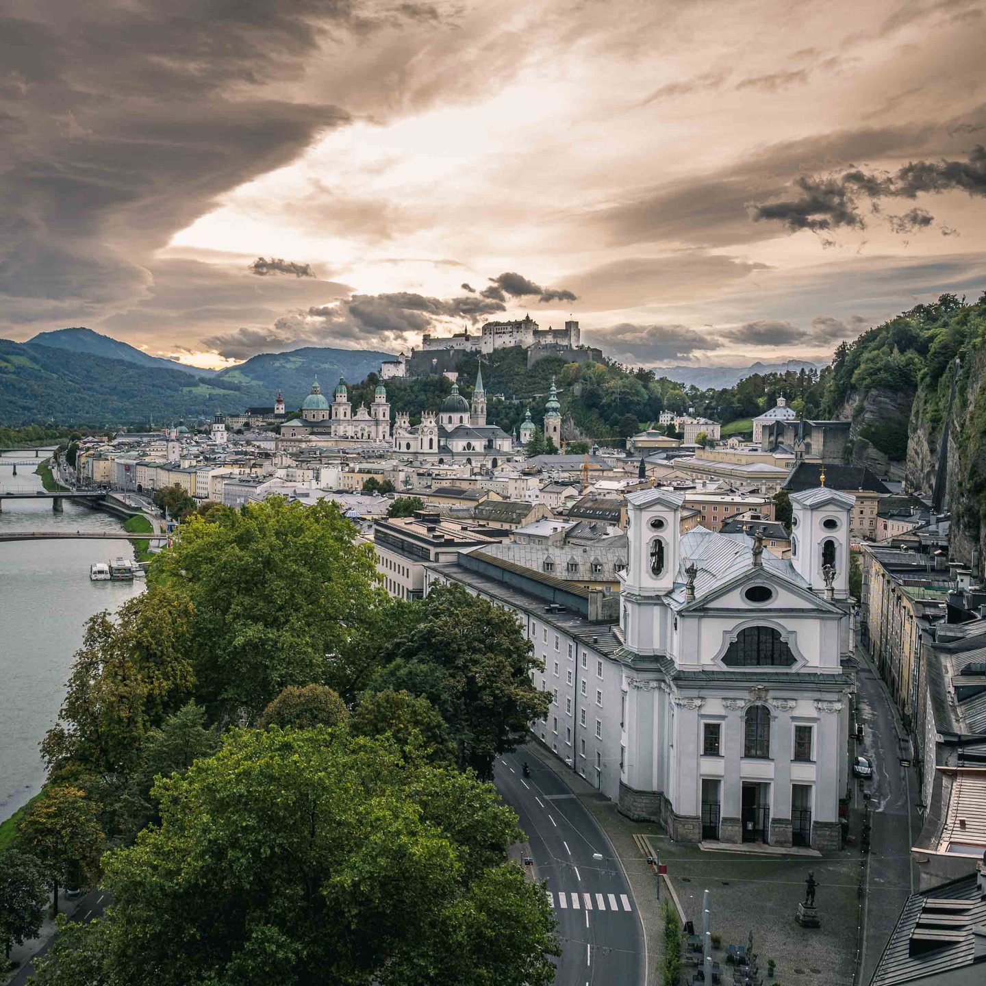 Wide view from Humboldtterrasse at dusk with the Markuskirche facade in the foreground and Hohensalzburg Fortress on the hill behind