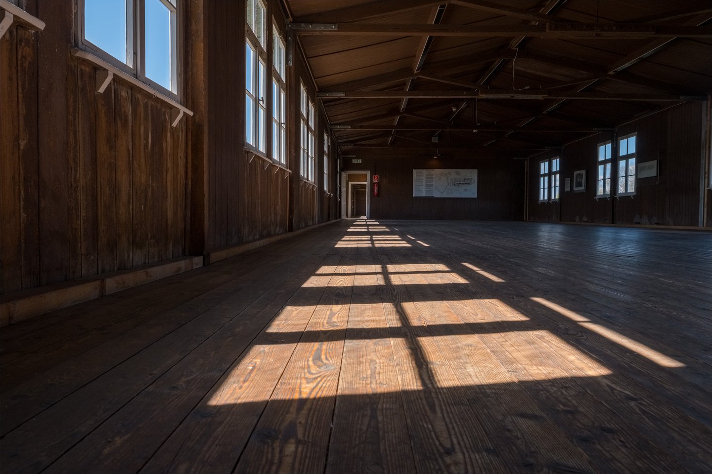 Sunlight from a row of windows falls in bright rectangles across the bare plank floor of an empty wooden prisoner barracks at the Mauthausen Memorial.