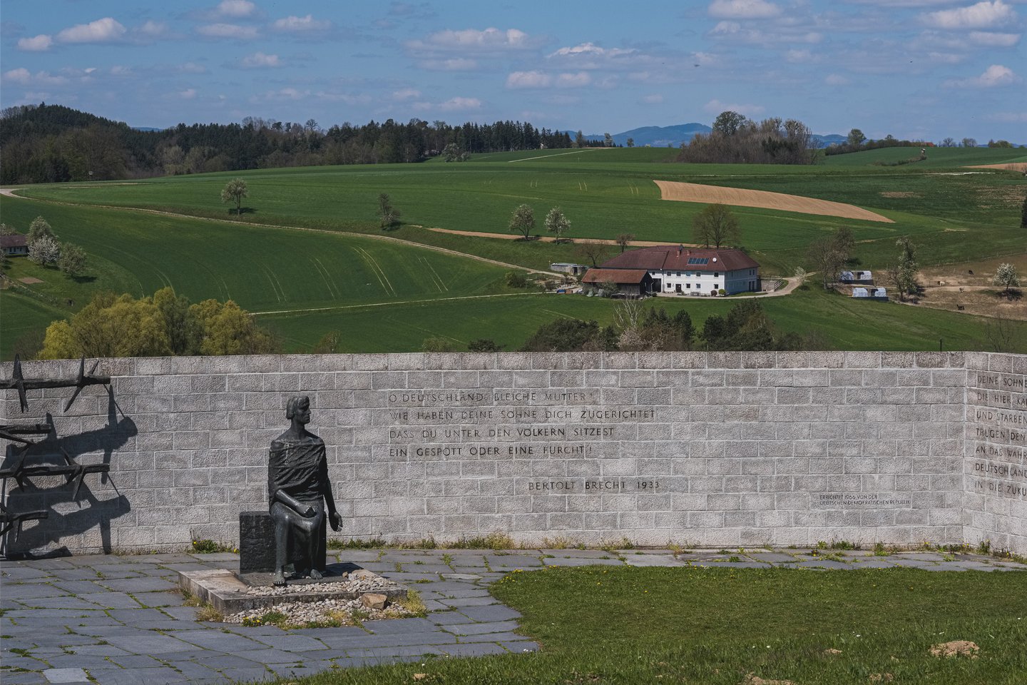 The long curved granite wall of the German national memorial at Mauthausen, carved with a Bertolt Brecht verse, with the bronze seated figure at its left end and farmland rising behind.