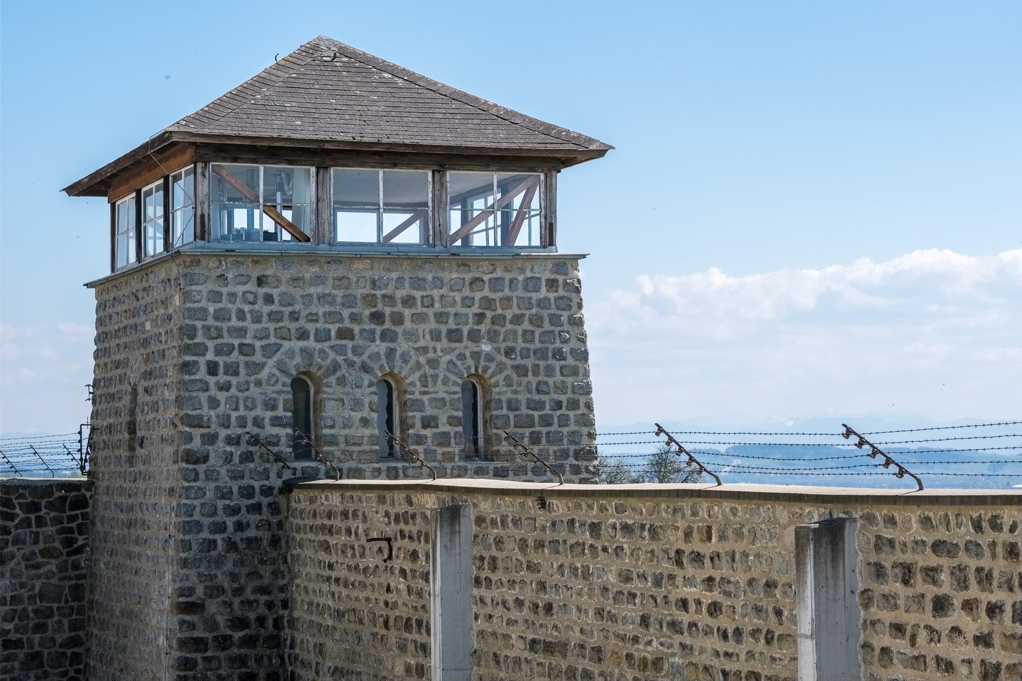 A stone guard tower with a timber and glass cabin rises above the barbed wire topped granite perimeter wall of the Mauthausen Memorial under a blue spring sky.