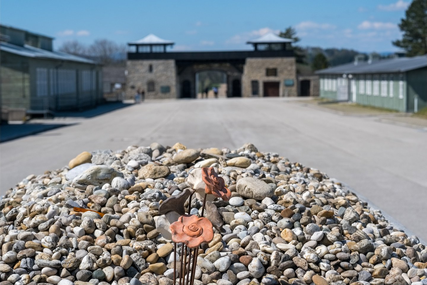 A mound of pebbles left as remembrance stones, with ceramic roses pushed into it, in front of the twin-towered stone gatehouse of the Mauthausen Memorial.