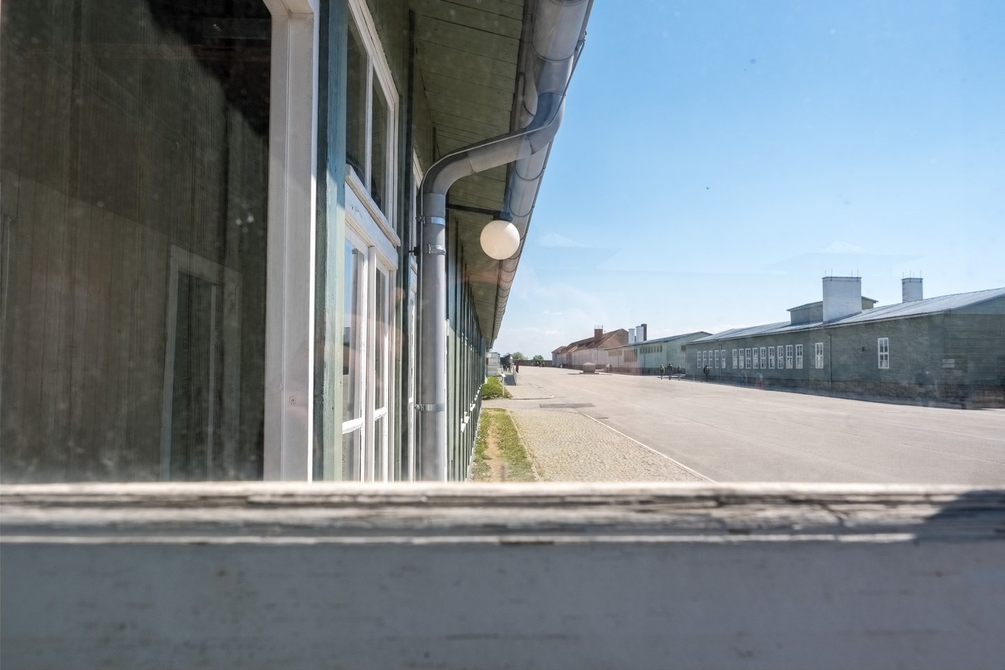 Seen through a barrack window at the Mauthausen Memorial, the long roll call square runs between rows of green timber barracks under a clear blue sky.