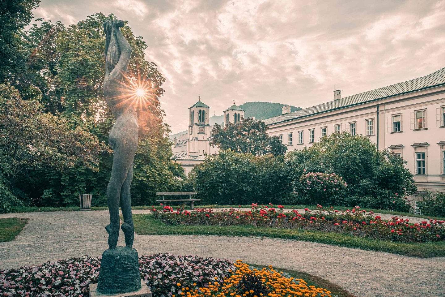 Sun flare behind a bronze figure in Mirabell Gardens with Andraekirche twin towers beyond in Salzburg.