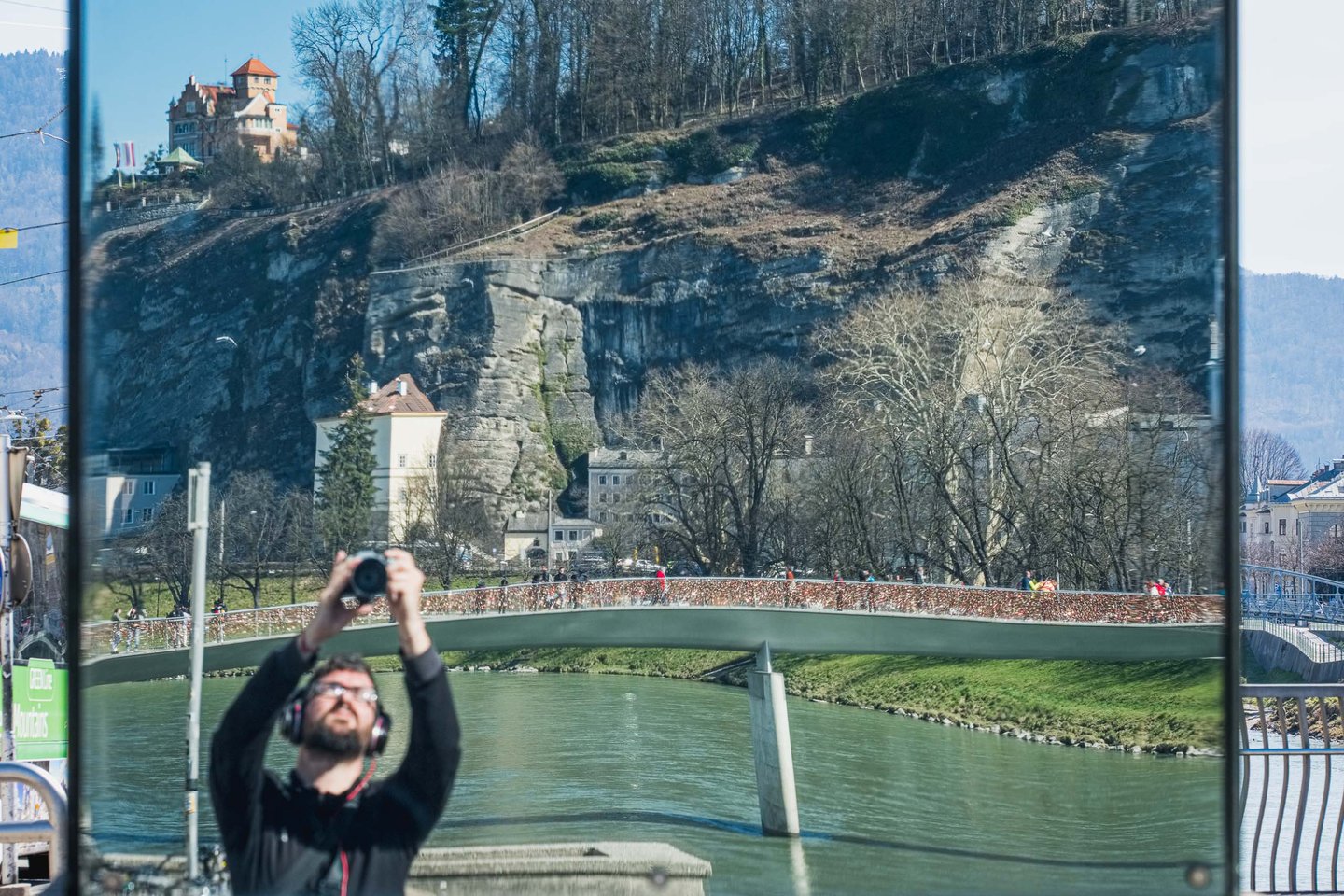 Reflection shows Makartsteg footbridge and the Moenchsberg cliff with a photographer at the Salzach in Salzburg.