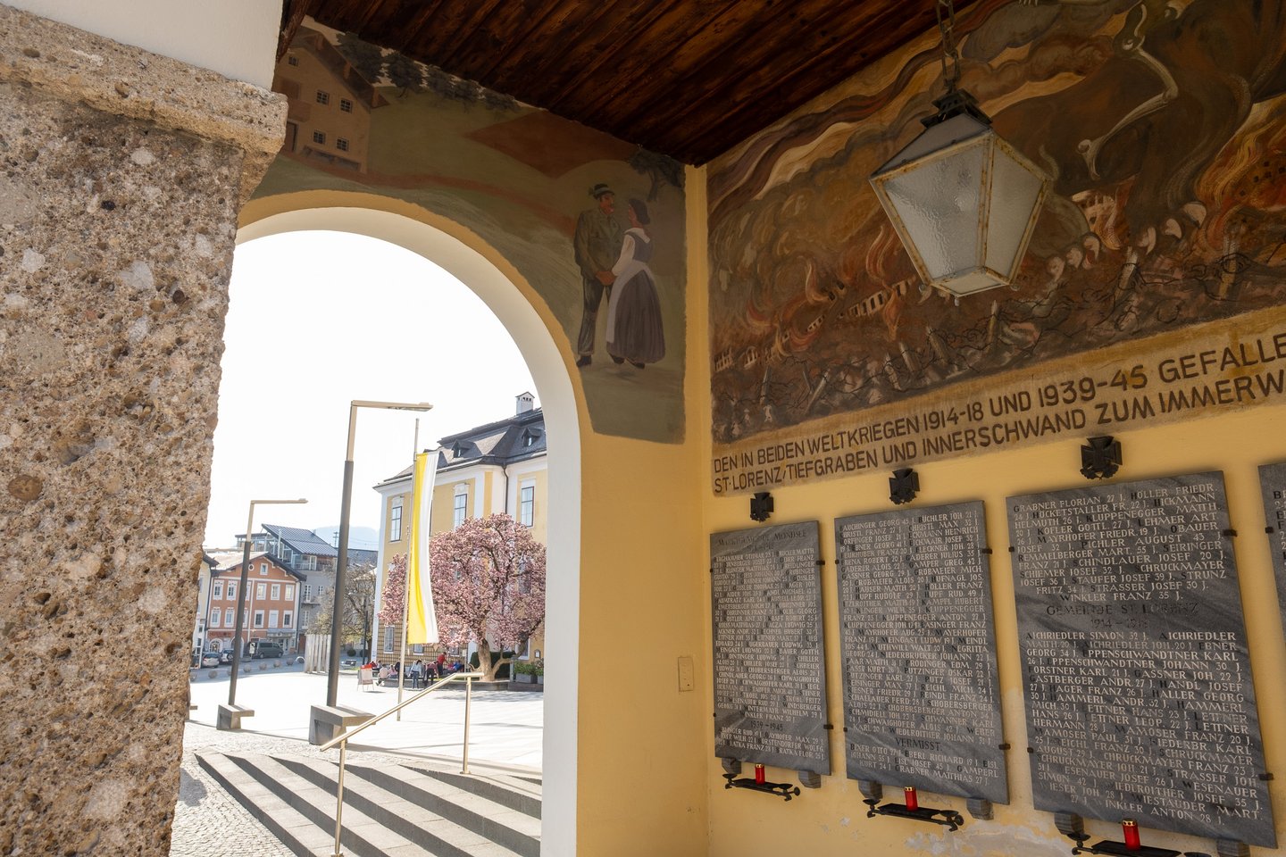 The war memorial arcade beside the Mondsee basilica, its wall painted with a battle scene above dark stone plaques listing the dead of both world wars, with the market square and a blossoming magnolia visible through the arch.