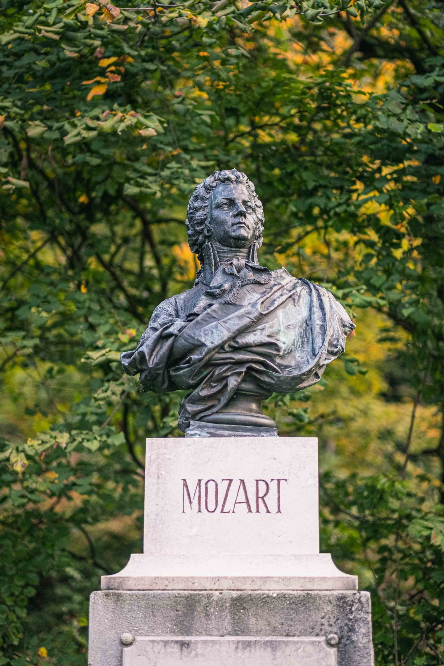 Bronze Mozart bust on a stone pedestal labelled MOZART among trees on Kapuzinerberg in Salzburg.