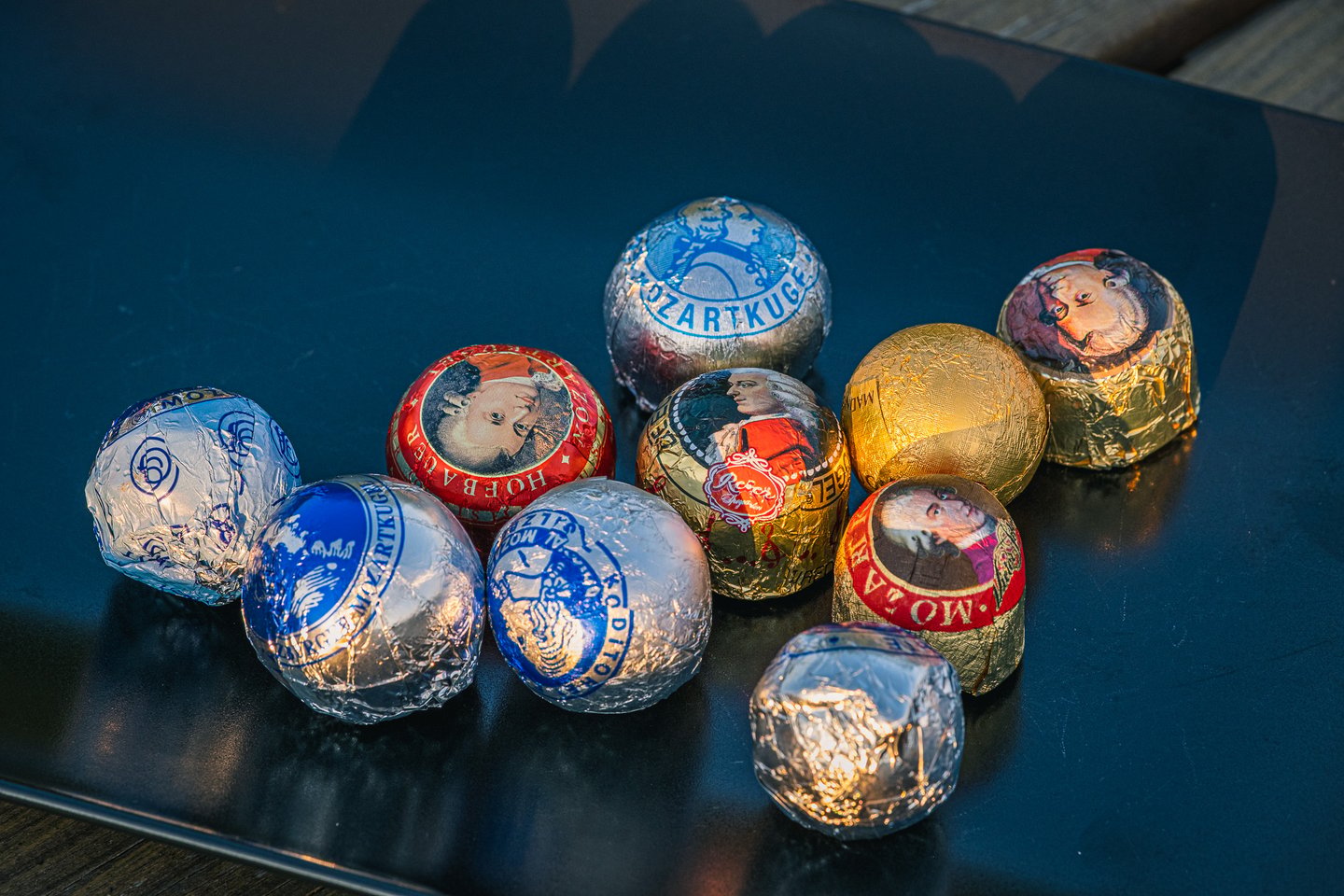 Ten Mozartkugeln from different makers, wrapped in silver, blue, red and gold foil with Mozart portraits, lie grouped on a dark table in low evening light.