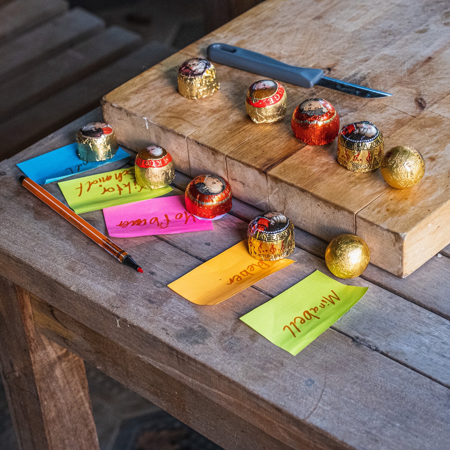 Eight foil-wrapped Mozartkugeln from different makers laid out on a wooden chopping board with handwritten coloured paper slips naming each brand.