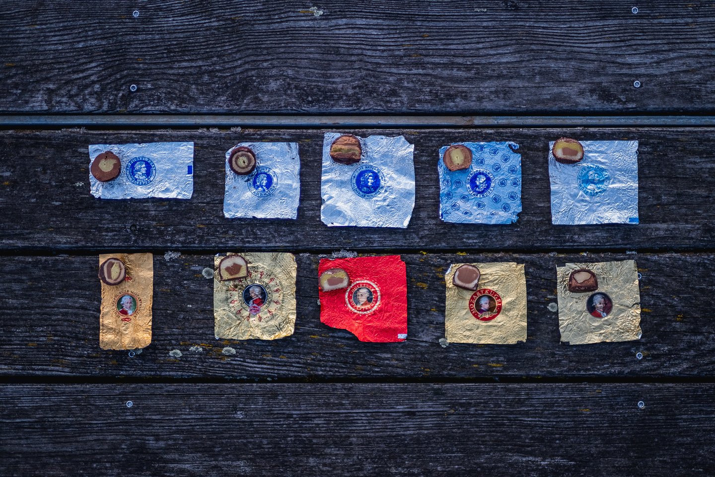Ten halved Mozartkugeln, each resting on its own foil wrapper, laid out in two rows on grey weathered planks, five silver and blue above and five gold and red below.