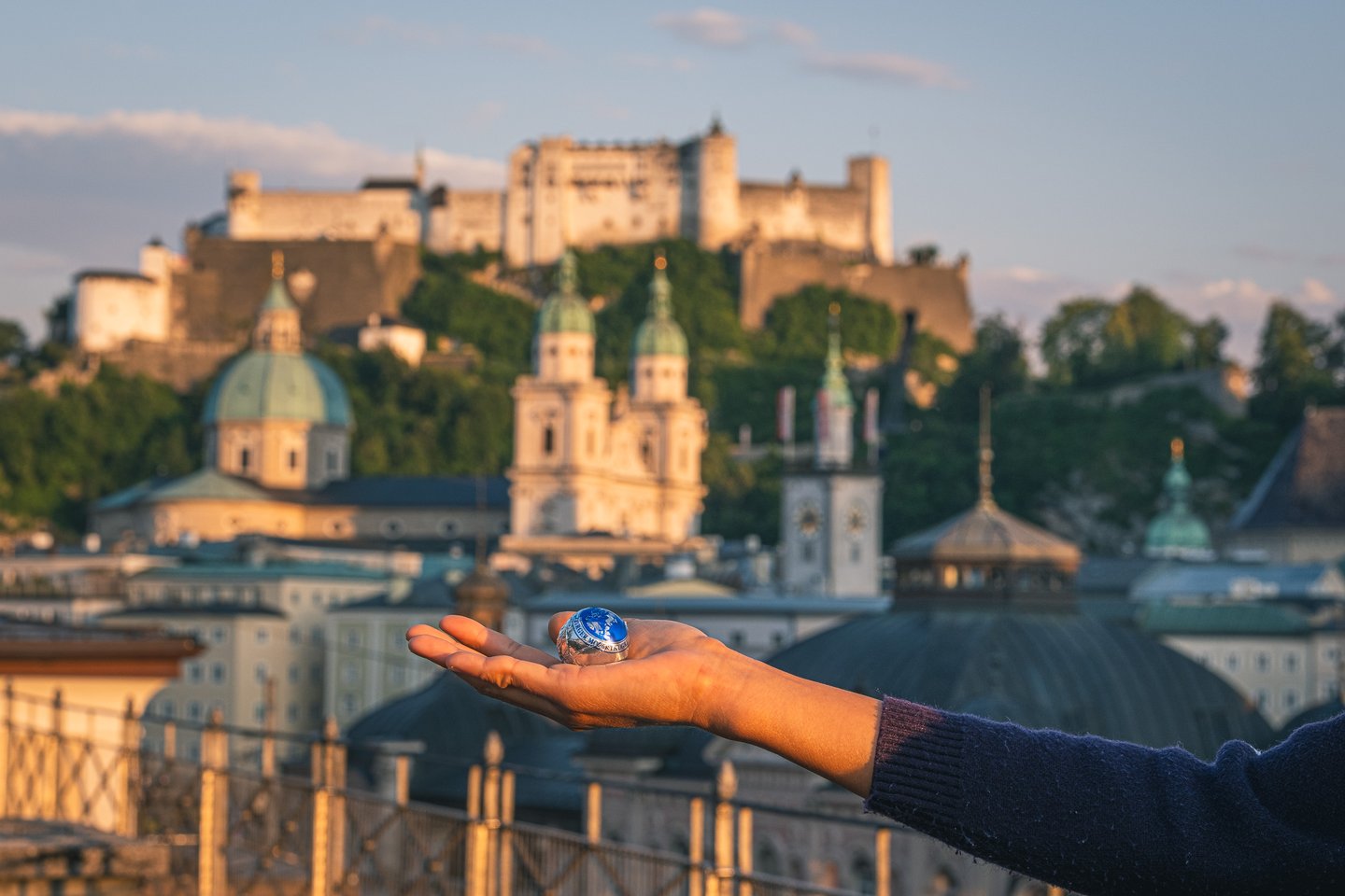 An open palm holds a single blue-foil Mozartkugel with the Hohensalzburg Fortress and the domes of Salzburg Cathedral lit gold behind it.