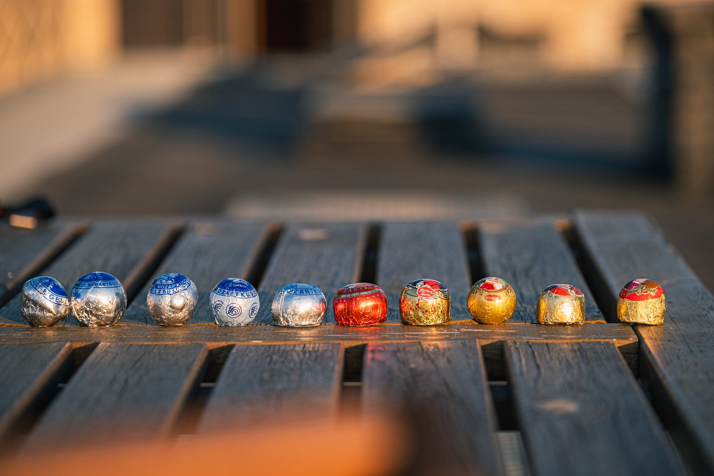 Ten Mozartkugeln from different makers lined up in a single row across the slats of a wooden deck table in low evening sun.