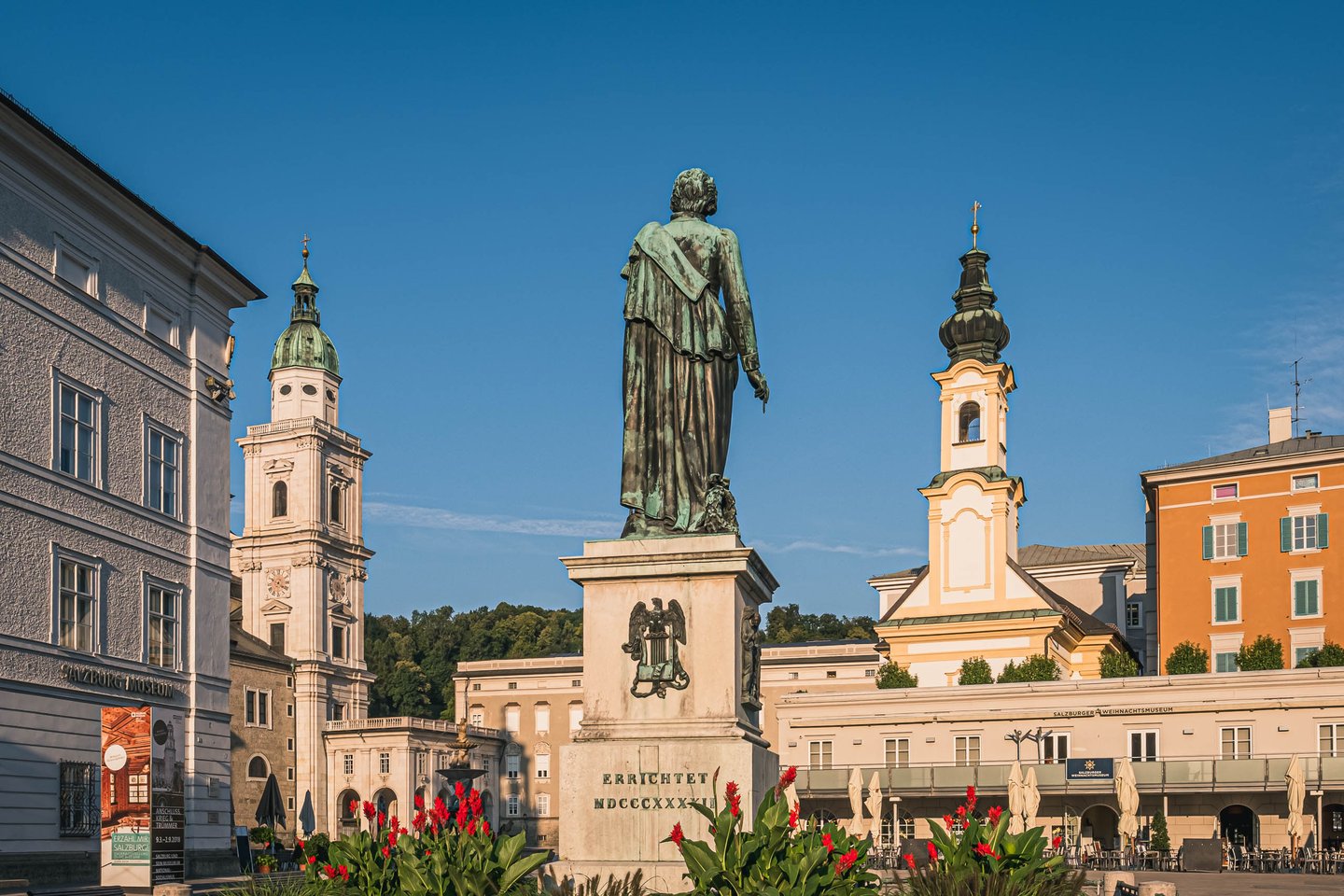 Bronze Mozart statue back view on Mozartplatz flanked by cathedral tower and Michaelskirche in Salzburg.