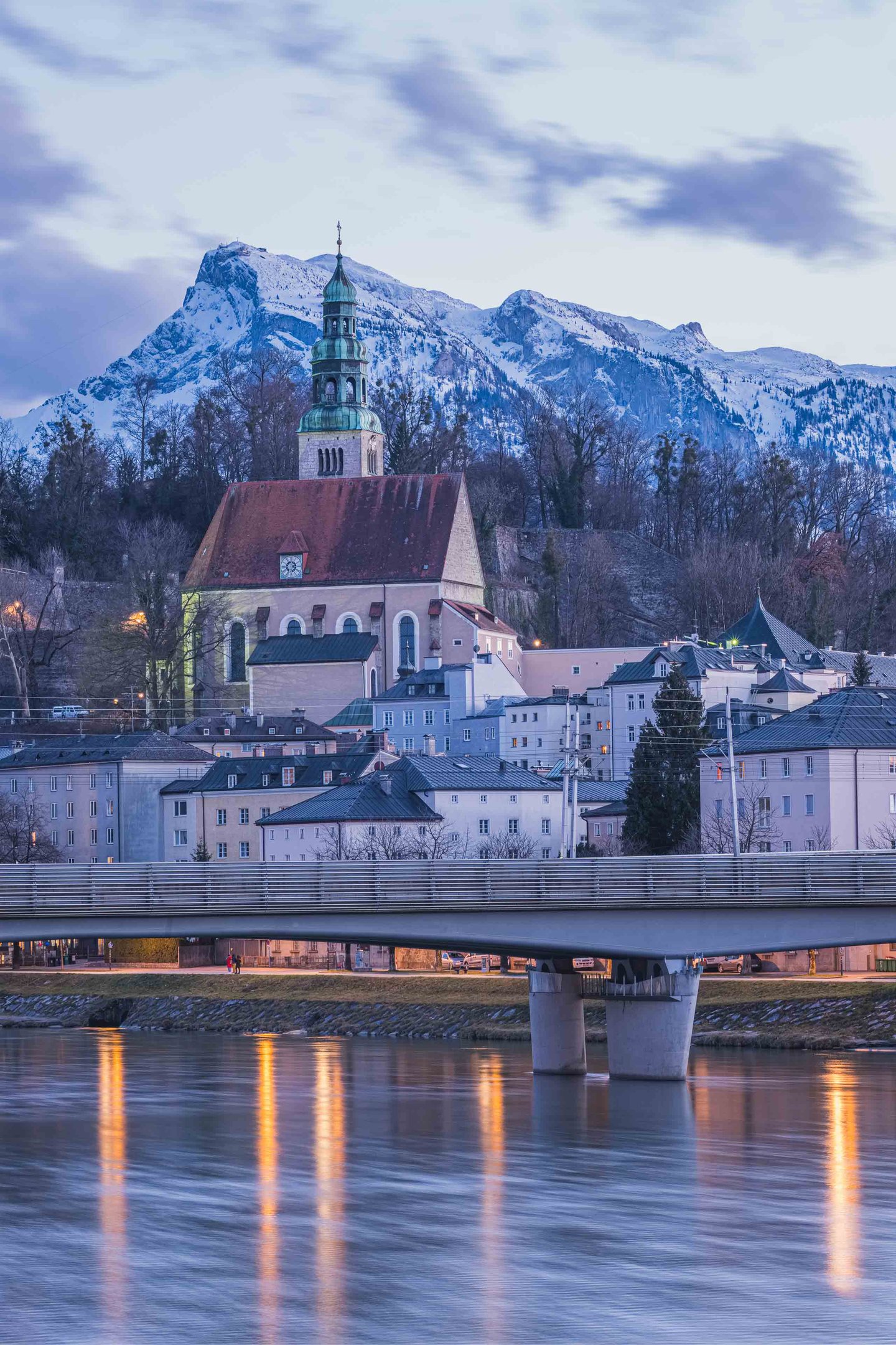 Mülln Church tower glows at dusk with Untersberg and a footbridge over the Salzach in Salzburg.