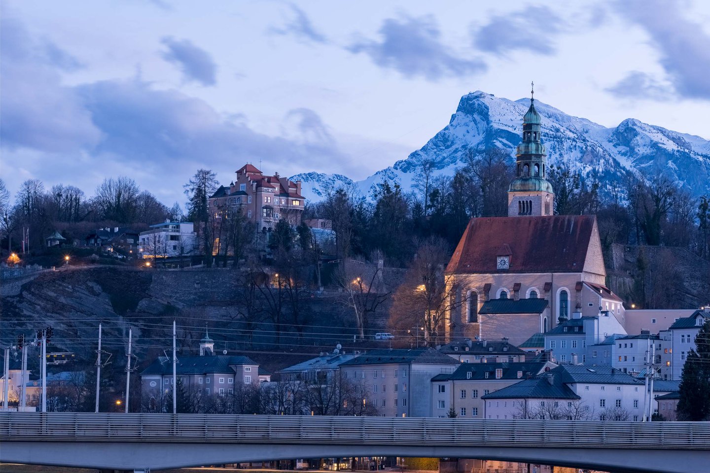 Mülln Church and hillside villas glow at dusk with Untersberg rising behind in Salzburg.