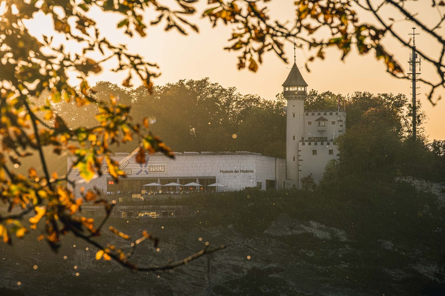 Museum der Moderne and its tower on the Mönchsberg glow at sunset through autumn leaves in Salzburg.