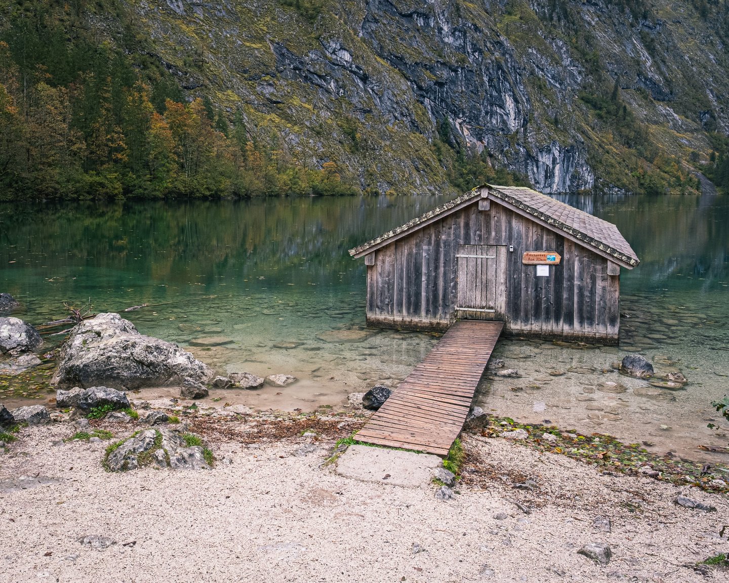 The Obersee boathouse seen across clear turquoise shallows from the pale gravel shore, with autumn woods and grey rock rising behind the lake.