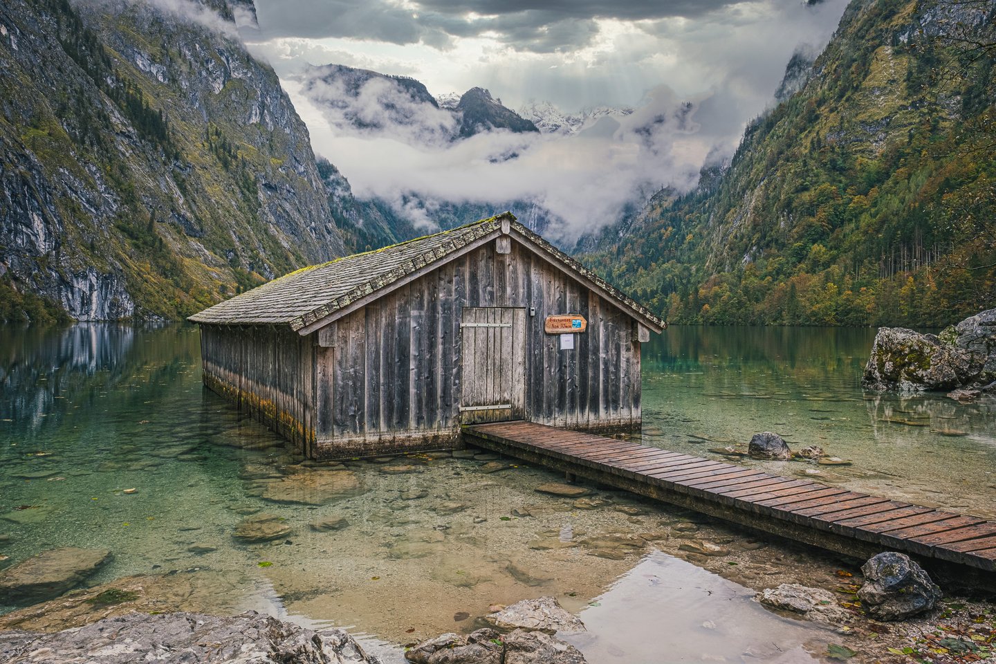 A three quarter view of the weathered wooden Obersee boathouse standing in glassy shallow water, with cloud filling the valley and snow on the far peaks.