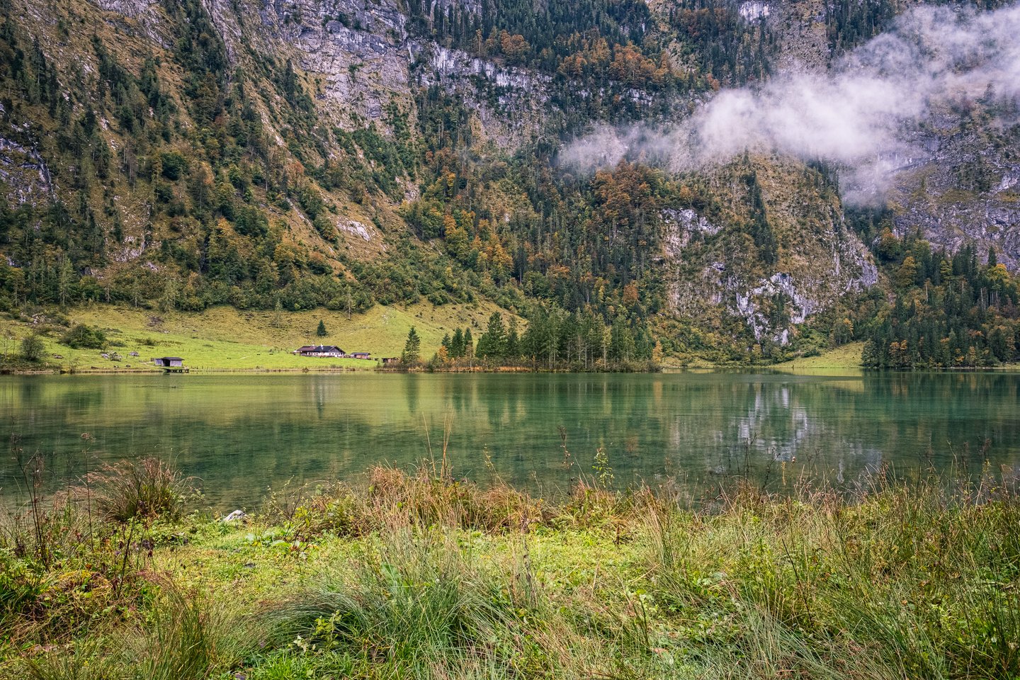 Across the still green water of the Obersee, the white Fischunkelalm farmhouse and its timber boat shed sit on a bright alp meadow under a huge grey cliff wall.