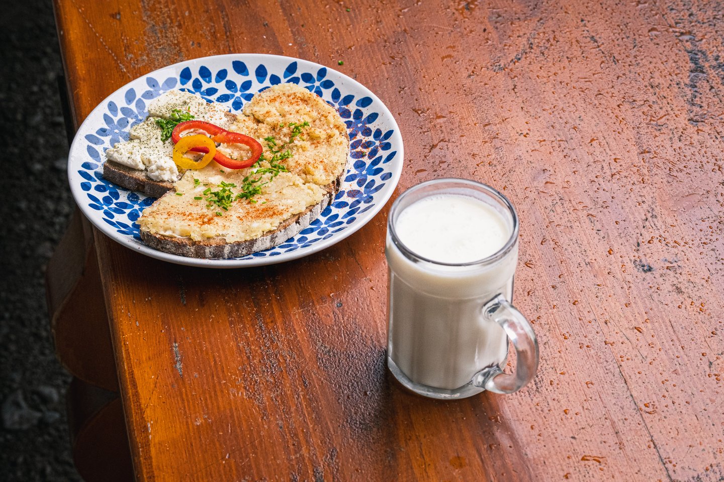 A plate of dark bread spread with cheese and garnished with chives and pepper rings sits beside a glass mug of fresh milk on a wet varnished bench.