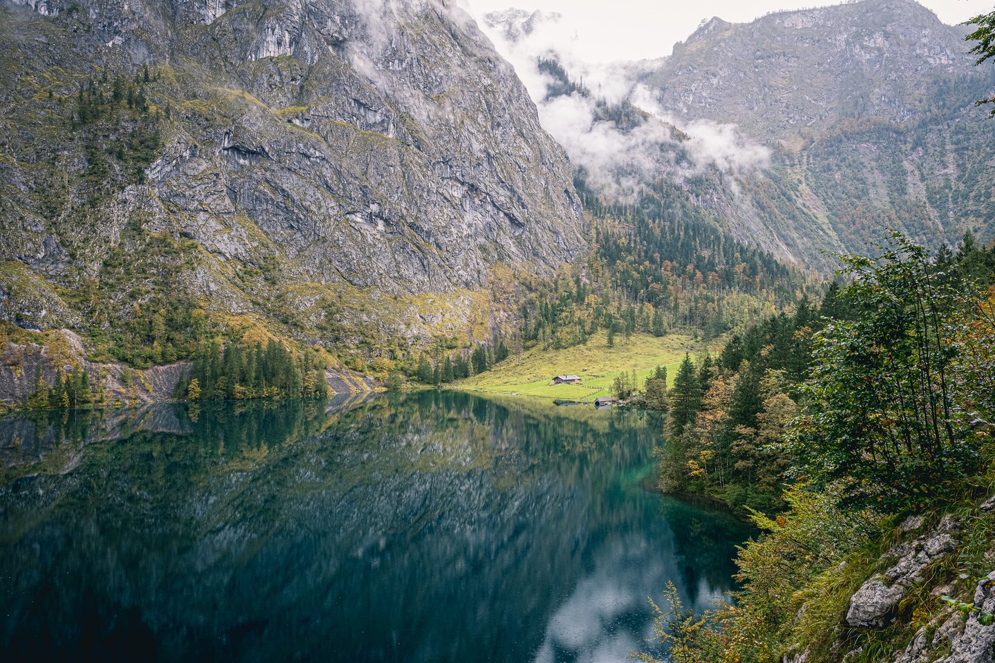 An open high view along the Obersee to the Fischunkelalm on its meadow, the grey cliff wall mirrored in the dark green water below.