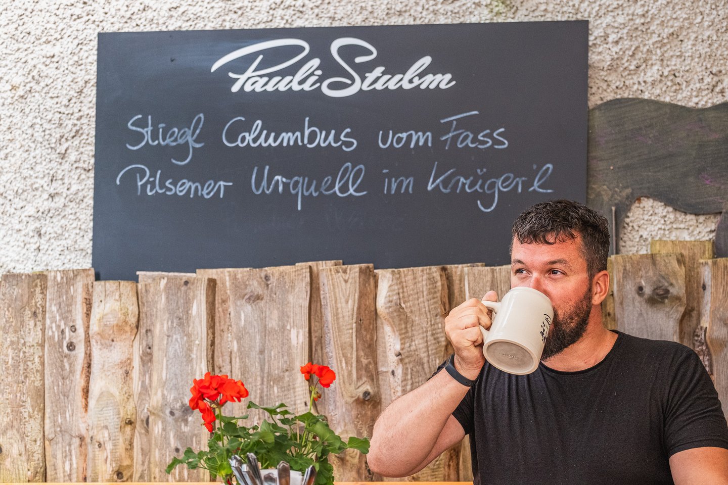 A bearded man in a black t-shirt drinks from a stoneware beer mug beneath the Pauli Stubm chalkboard offering Stiegl Columbus on tap and Pilsner Urquell in the Krügerl.