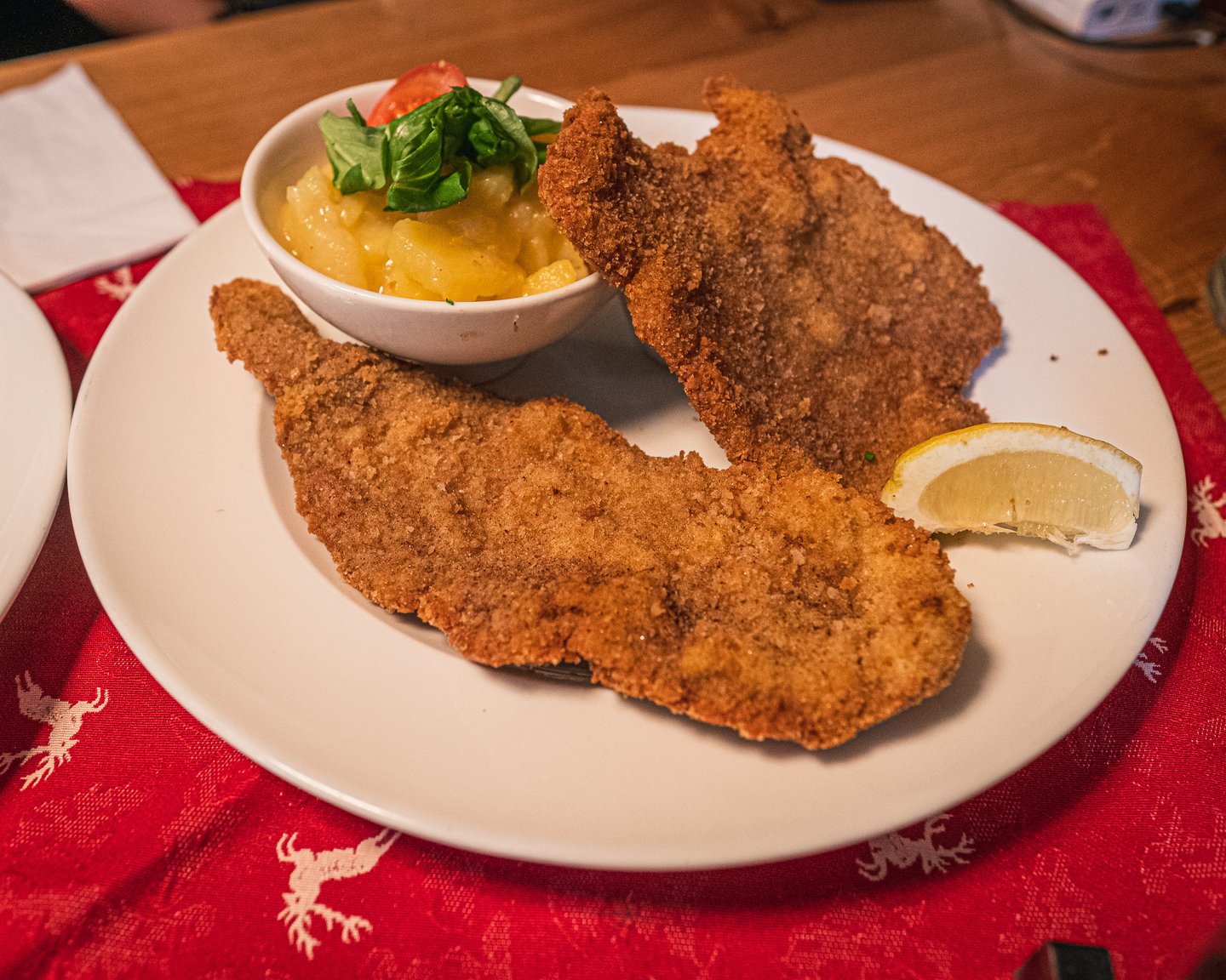Two breaded schnitzel on a white plate with a bowl of Austrian potato salad garnished with basil and tomato and a lemon wedge, on a red deer-patterned runner at Pauli Stubm in Salzburg.