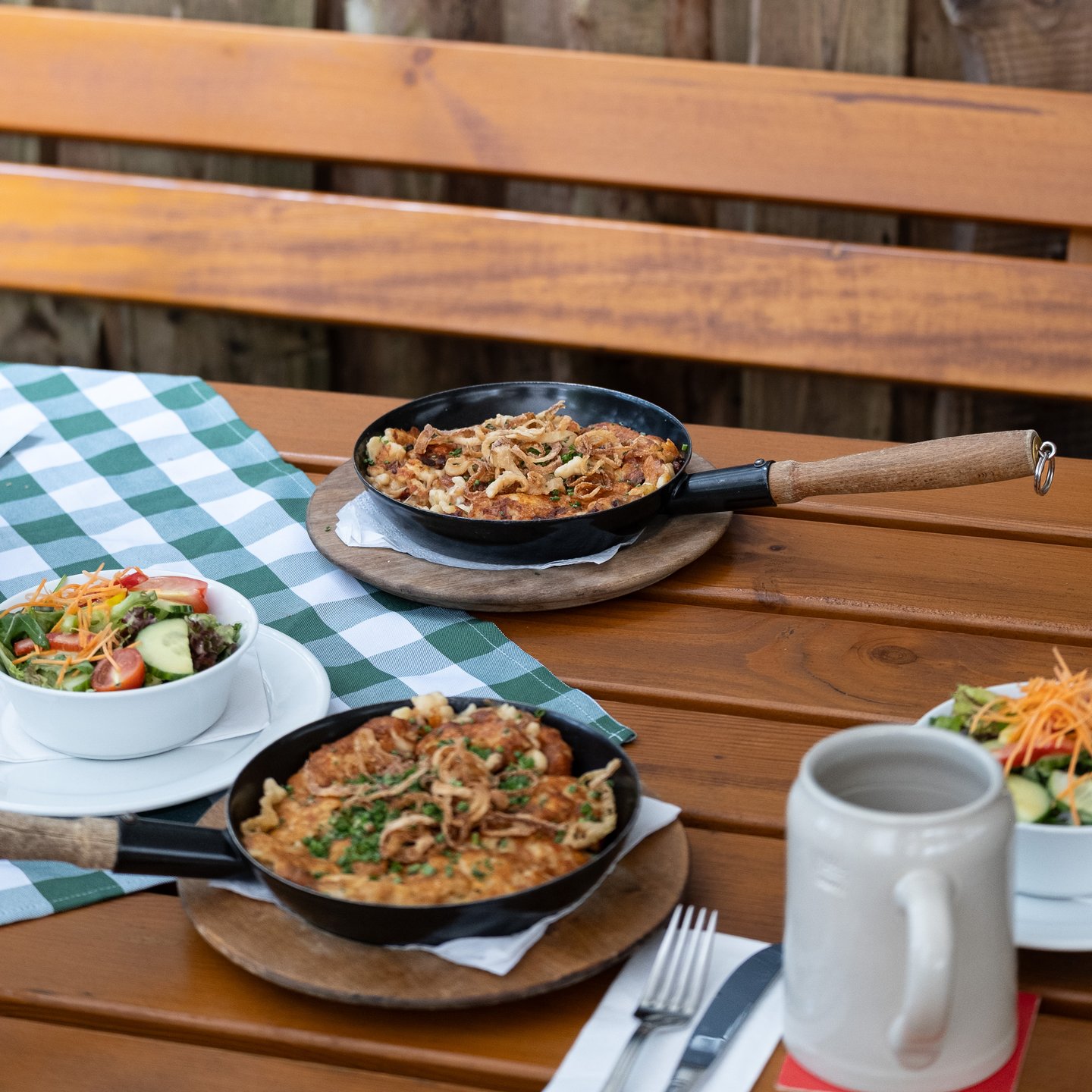 A beer garden table laid for two, with two pans of Kasnocken on wooden boards, two bowls of side salad, cutlery and a stoneware stein, in front of a wooden bench and fence.