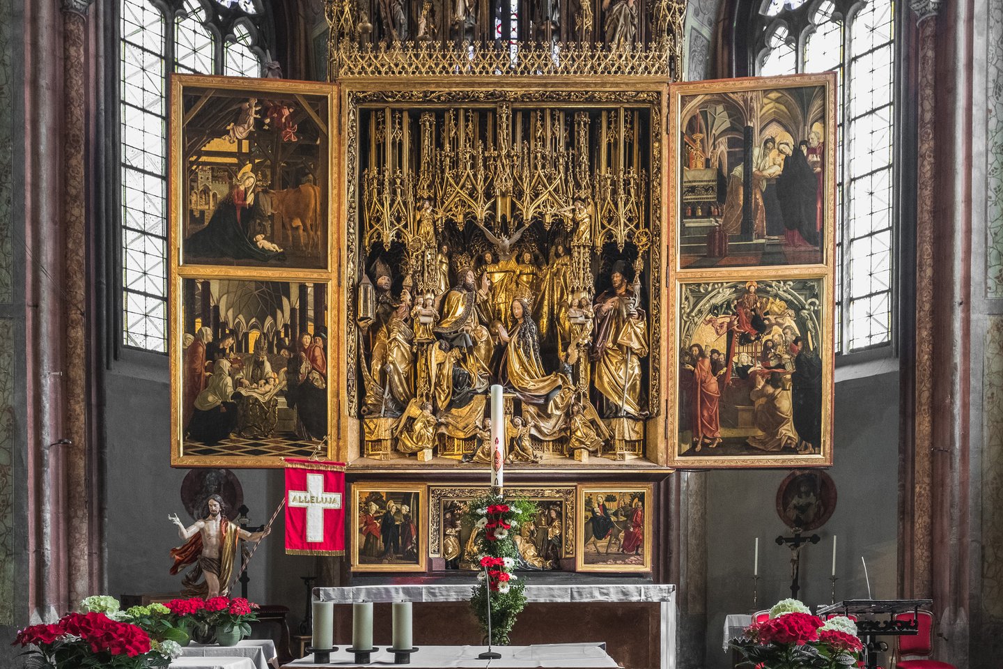 The Michael Pacher winged altarpiece in the church of St. Wolfgang stands fully open, its gilded Coronation of the Virgin shrine flanked by painted panels, with a red Alleluja banner, a paschal candle and a Risen Christ figure in front.