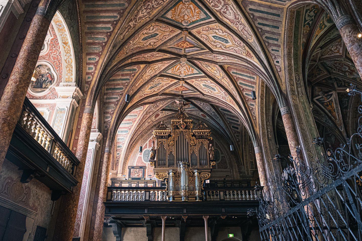 Looking up inside the pilgrimage church of St. Wolfgang at the richly painted rib vaulting in red, green and gold above the gilded organ on its wooden gallery.