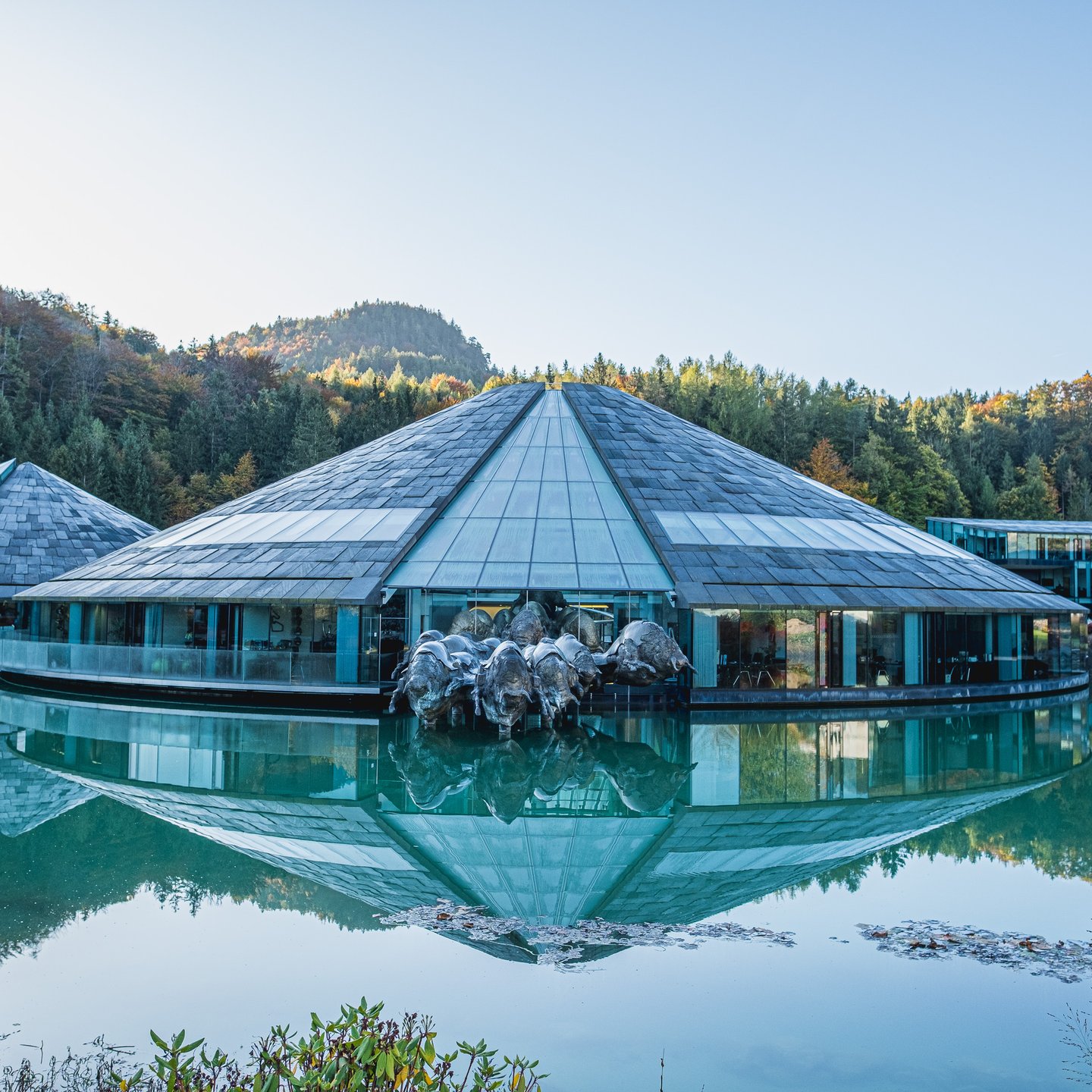 The main cone of the Red Bull headquarters seen head on, the bronze bull herd charging out over the water and the whole building doubled in the mirror-still lake.