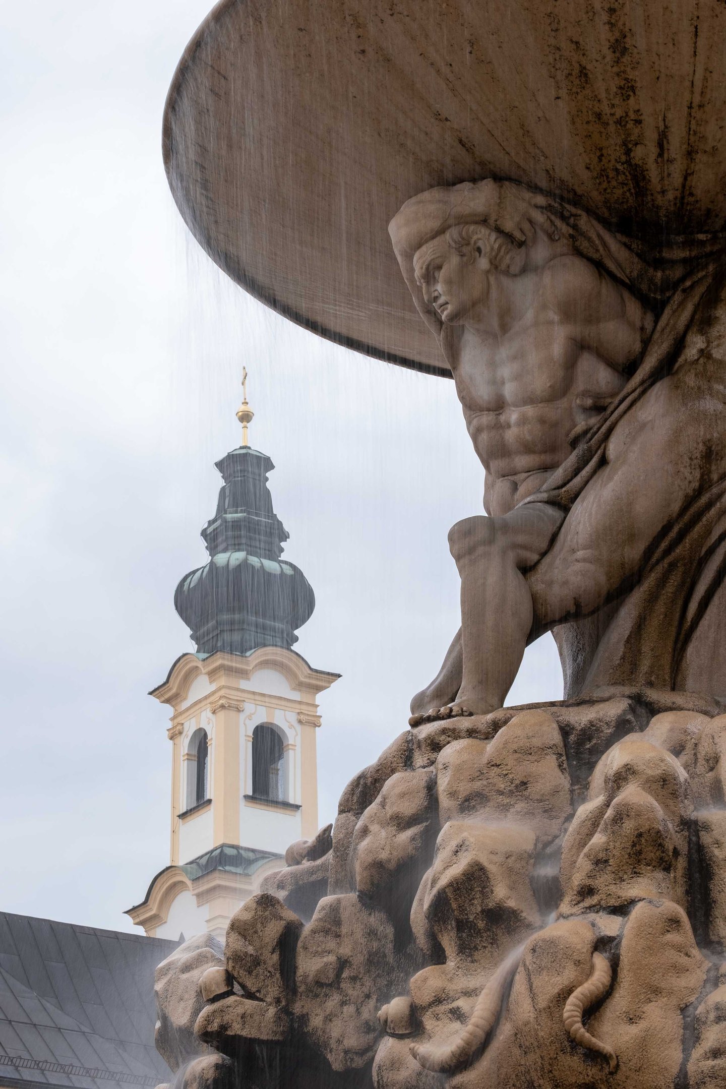 Atlas figure holding fountain basin on Residenzbrunnen with Michaelskirche tower behind in Salzburg