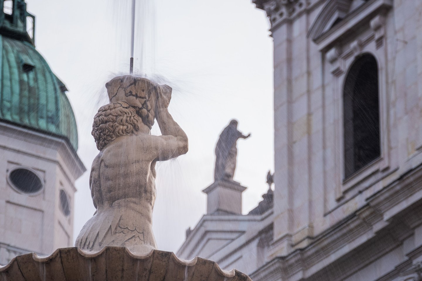 Triton figure of the Residenz Fountain spouts water with the Cathedral dome behind in Salzburg old town.