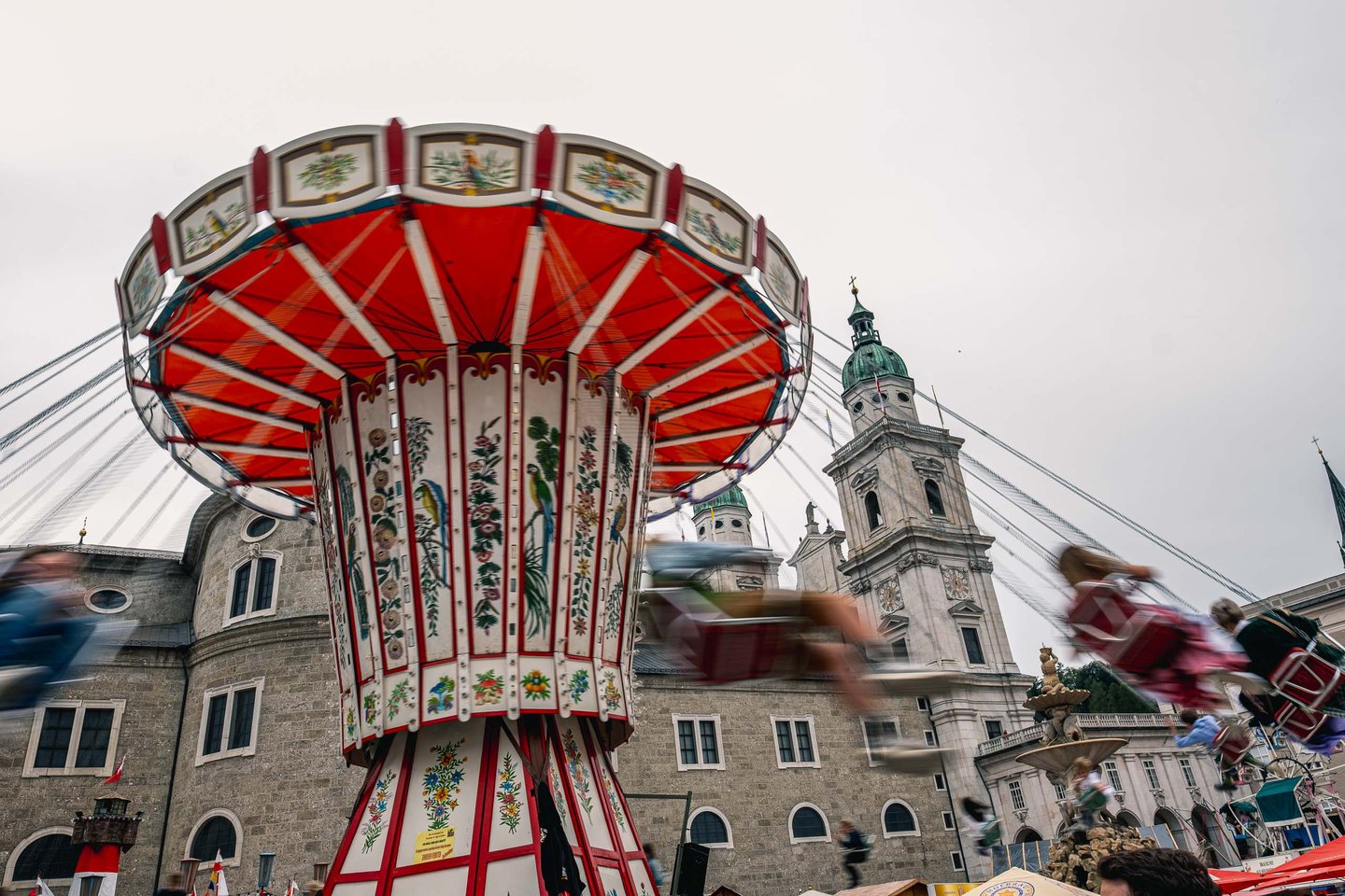 A painted chair-o-plane carousel spins at the Rupertikirtag funfair with the Salzburg Cathedral tower behind on Residenzplatz.