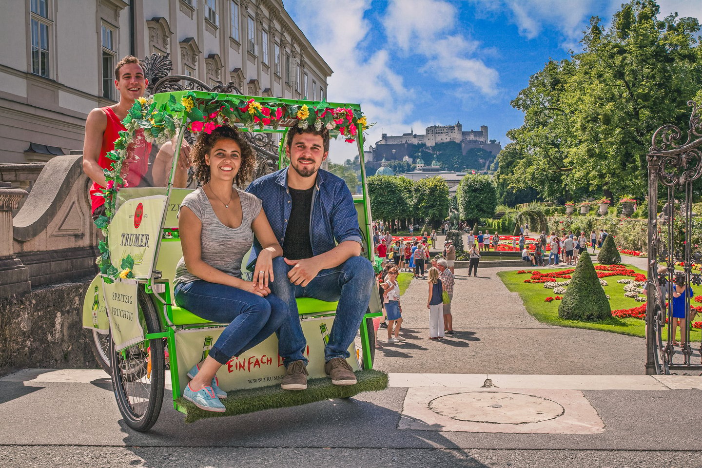 A couple sits in a flower-trimmed rickshaw at the wrought-iron gate of the Mirabell Gardens, the parterre beds and Hohensalzburg fortress framed behind them.