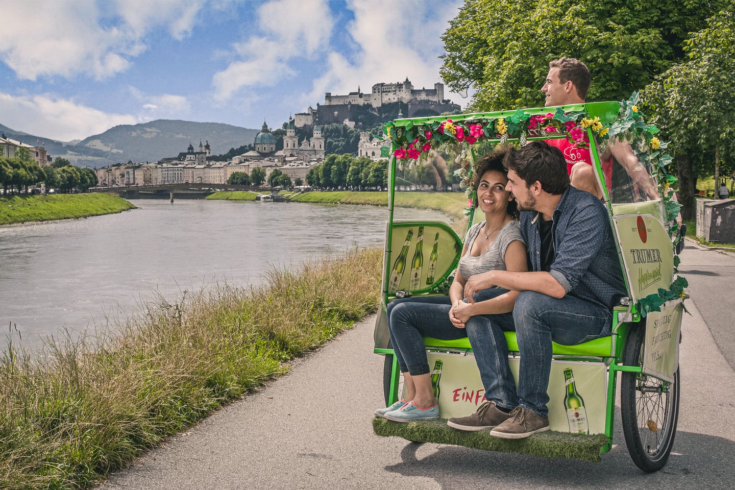 A couple rides in a flower-trimmed green rickshaw along the Salzach riverside path, with the old town domes and Hohensalzburg fortress on the far bank.
