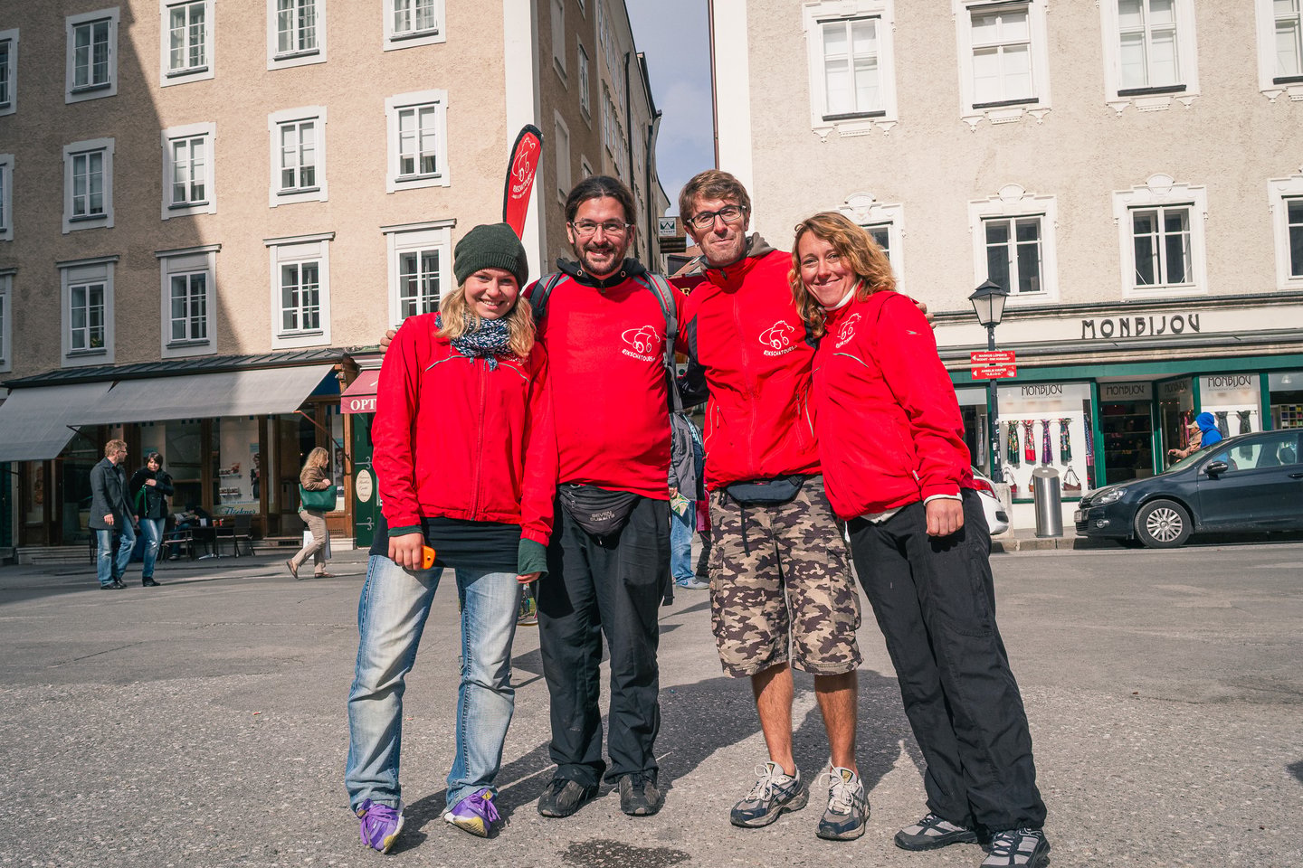 Four Rikschatours crew members in matching red jackets stand arm in arm on a paved old town square, a red teardrop banner with the company logo rising behind them.