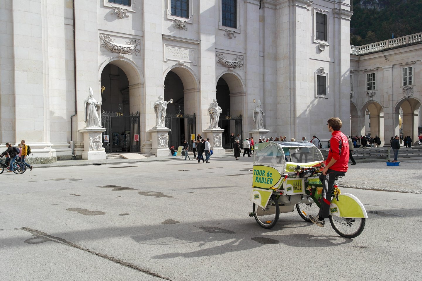 A driver pedals the lime green Sigl Tour de Radler pedicab across Domplatz, the white marble west facade of Salzburg Cathedral with its four saint statues and the dates 774, 1628 and 1959 rising behind.