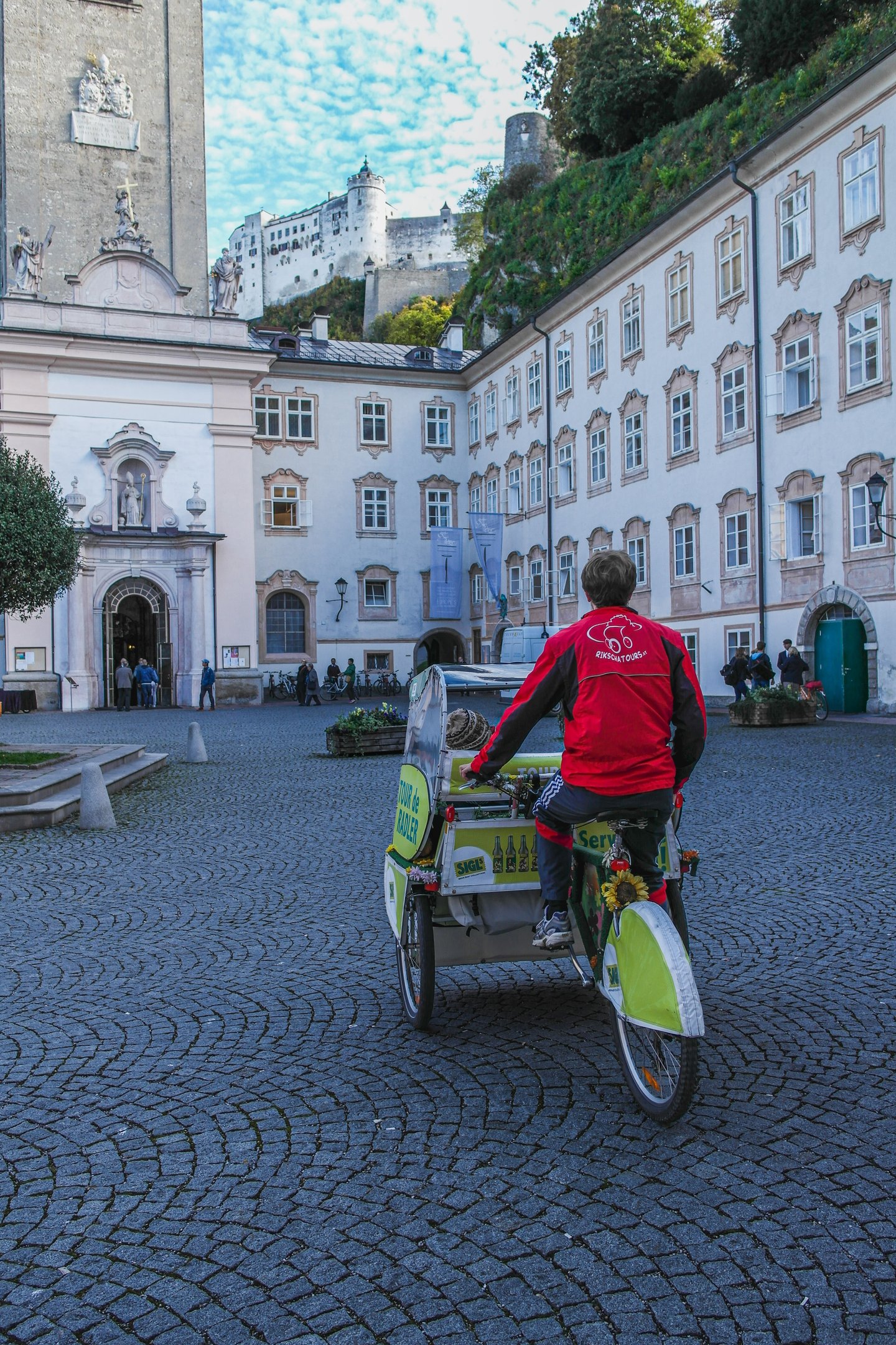 A driver in a red Rikschatours jacket rides the Sigl Tour de Radler pedicab across the cobbled courtyard of St Peter's Abbey, the church portal on the left and Hohensalzburg Fortress on the cliff above.