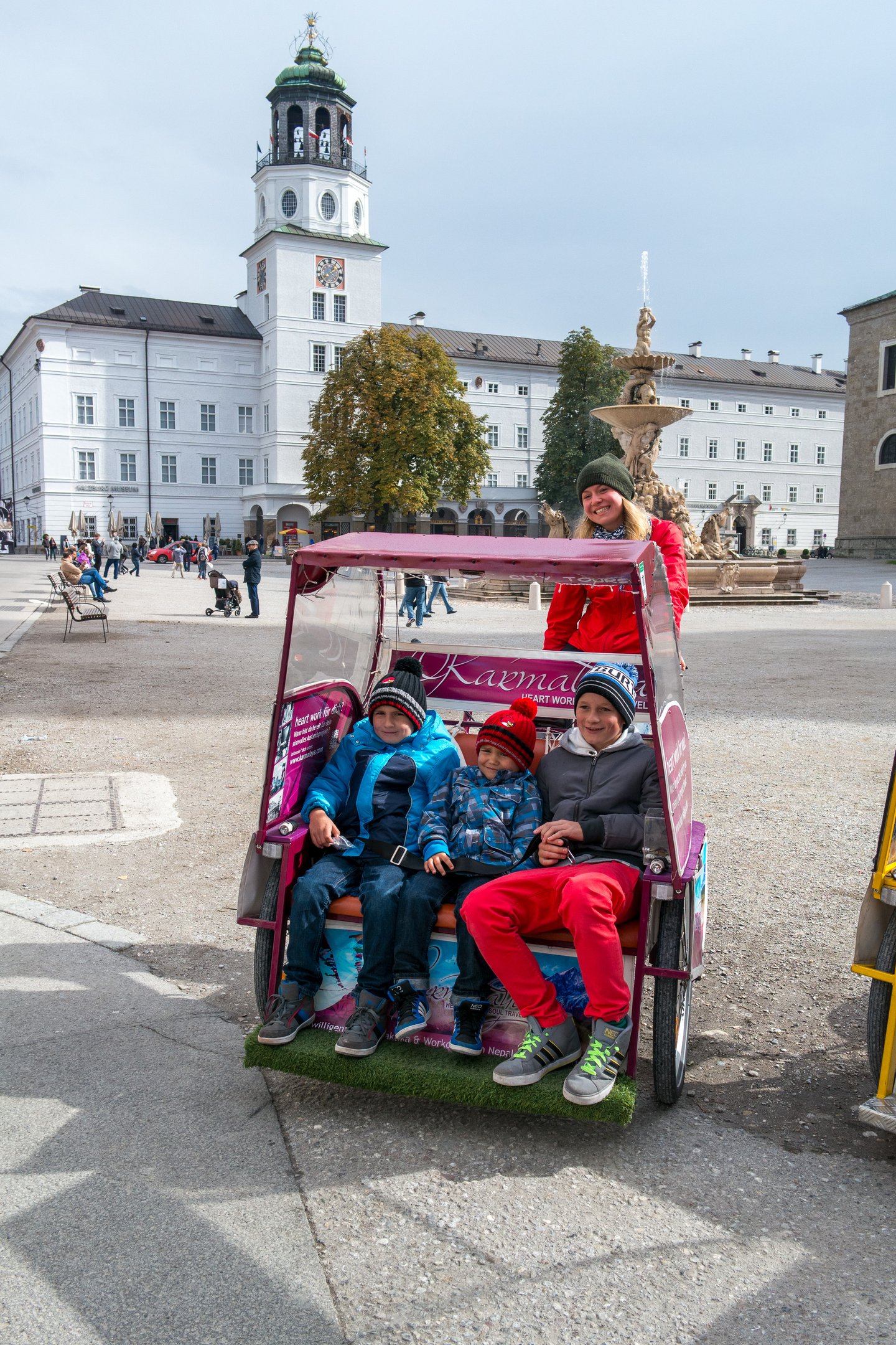 Three children in winter jackets and bobble hats sit belted into a pink Rikschatours pedicab on Residenzplatz, a smiling driver behind them and the Glockenspiel tower and Residenz fountain beyond.
