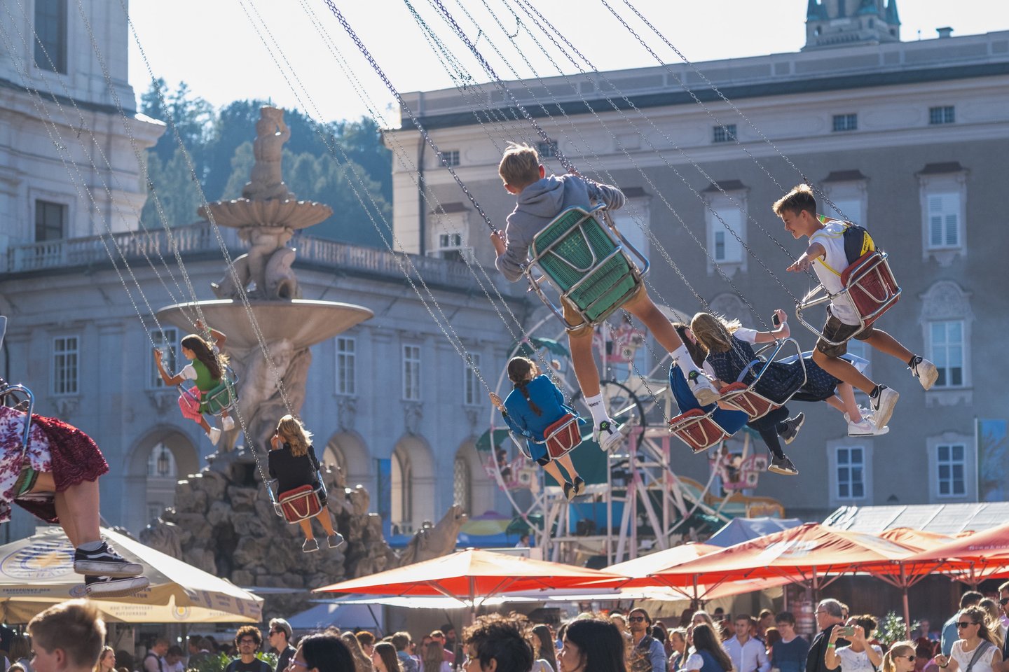 Children and teenagers fly outward on the chain swing carousel at the Rupertikirtag, with the Residenzbrunnen fountain and the Residenz facade behind them and beer garden umbrellas below.