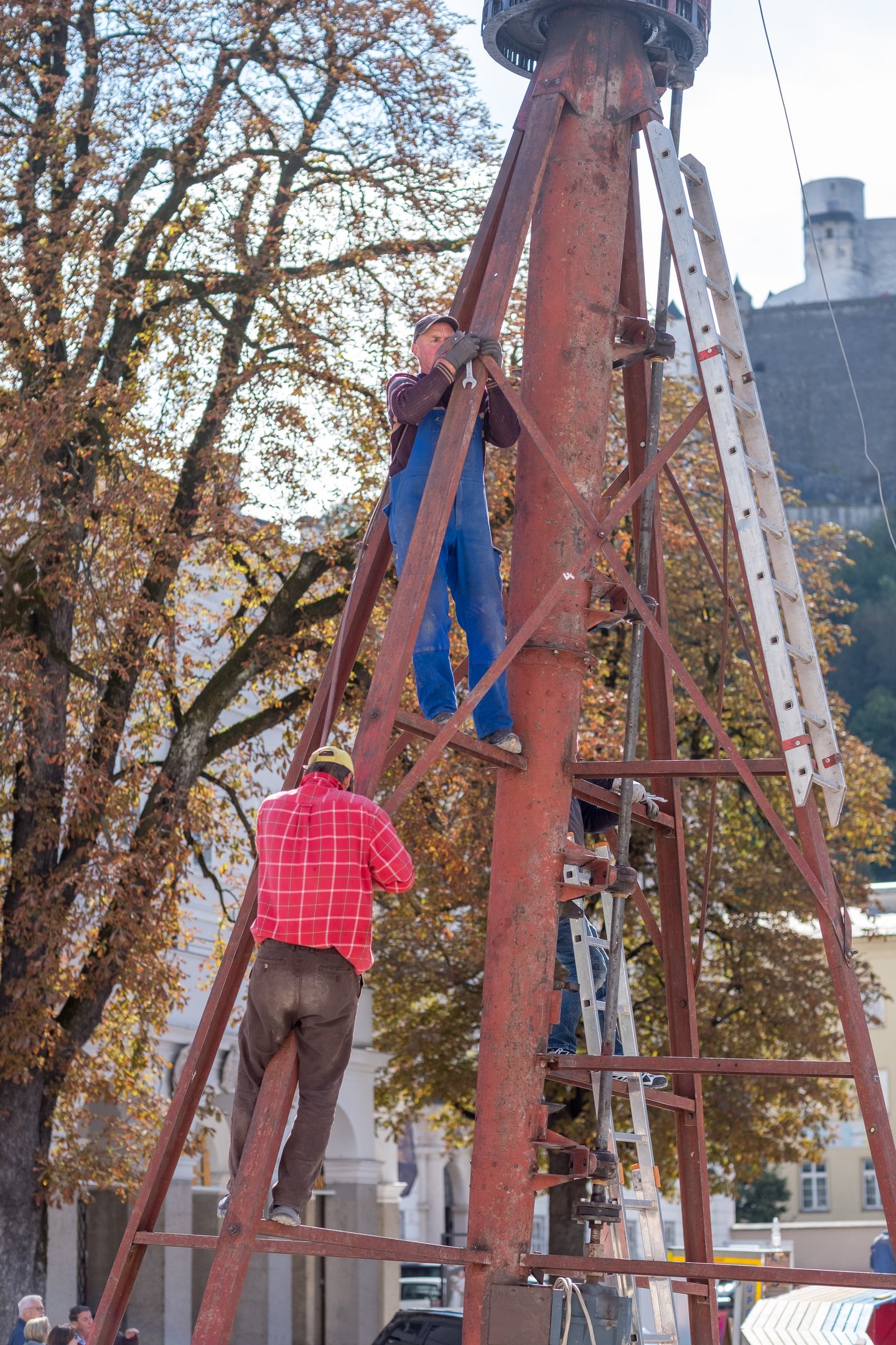 Workmen in blue overalls and a red checked shirt climb the rusty red lattice of a fairground ride being taken apart, with autumn chestnut trees, an arcaded white building and Hohensalzburg Fortress rising behind.