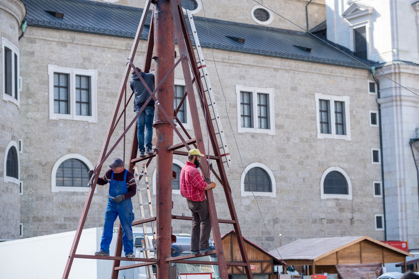 Three workmen stand on the rusty red steel frame of a fairground ride, unbolting it around the central column, with wooden market huts below them and a large stone Salzburg old town facade behind.