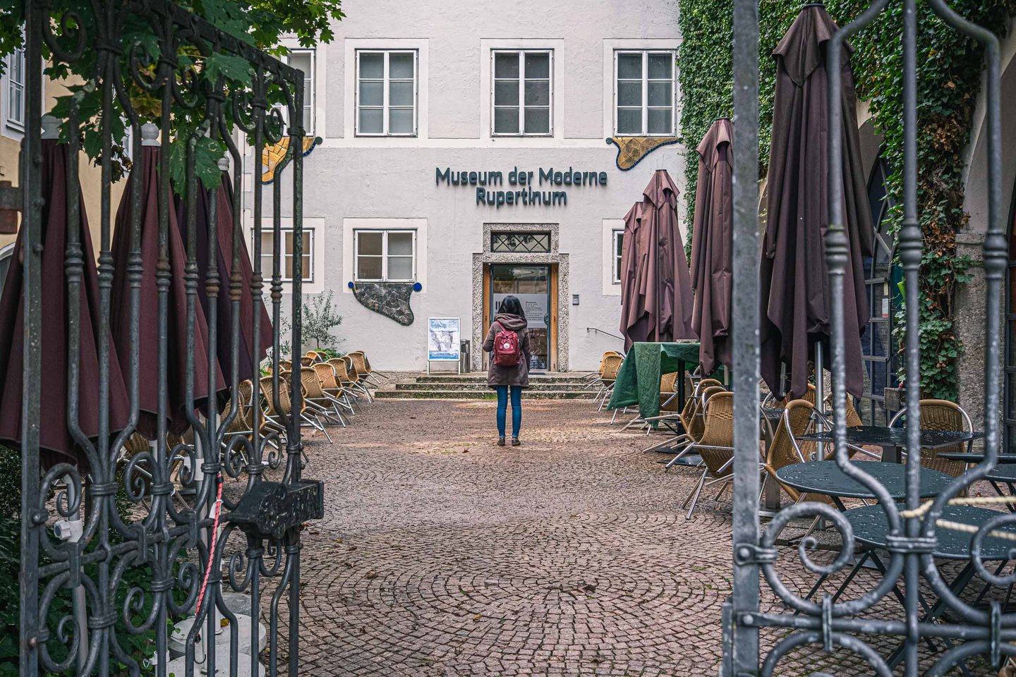 View through an iron gate toward the Museum der Moderne Rupertinum entrance with cafe chairs in Salzburg old town.