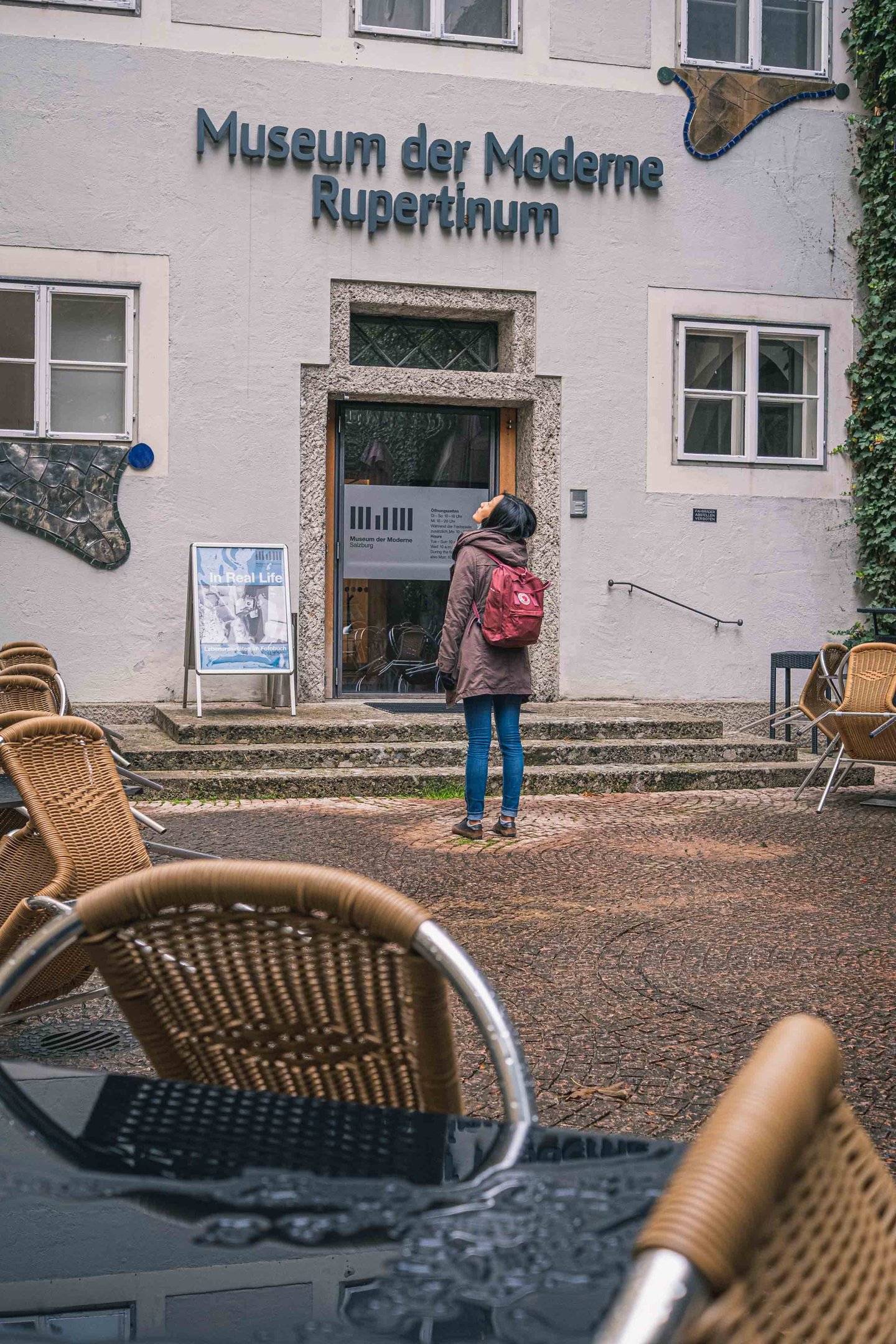 Woman in red backpack approaches the Museum der Moderne Rupertinum entrance framed by cafe chairs in Salzburg.