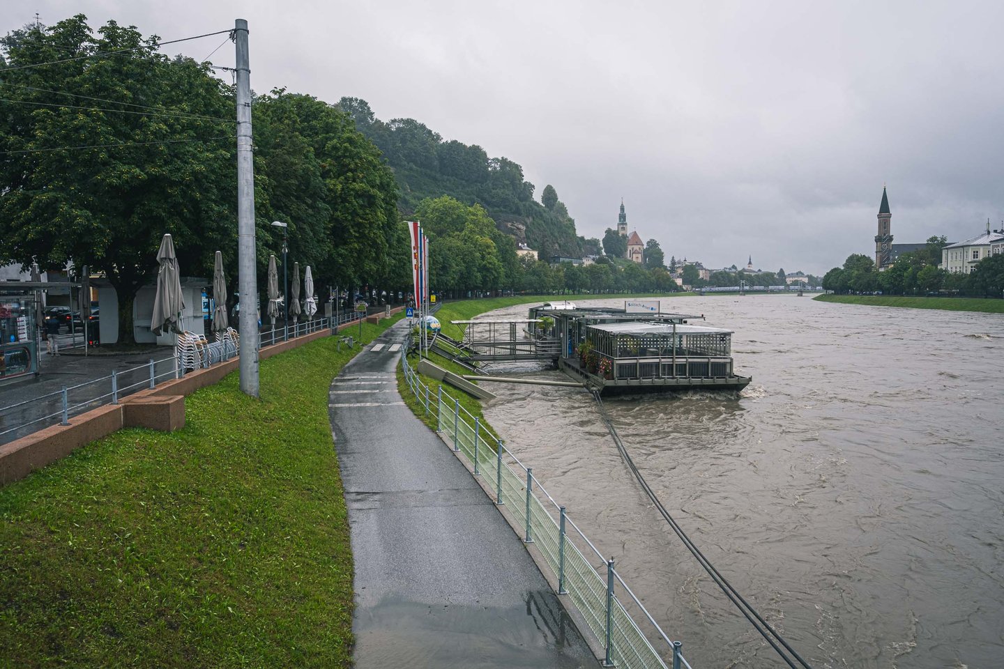 Flooded Salzach river along the promenade with Christuskirche tower visible in Salzburg.