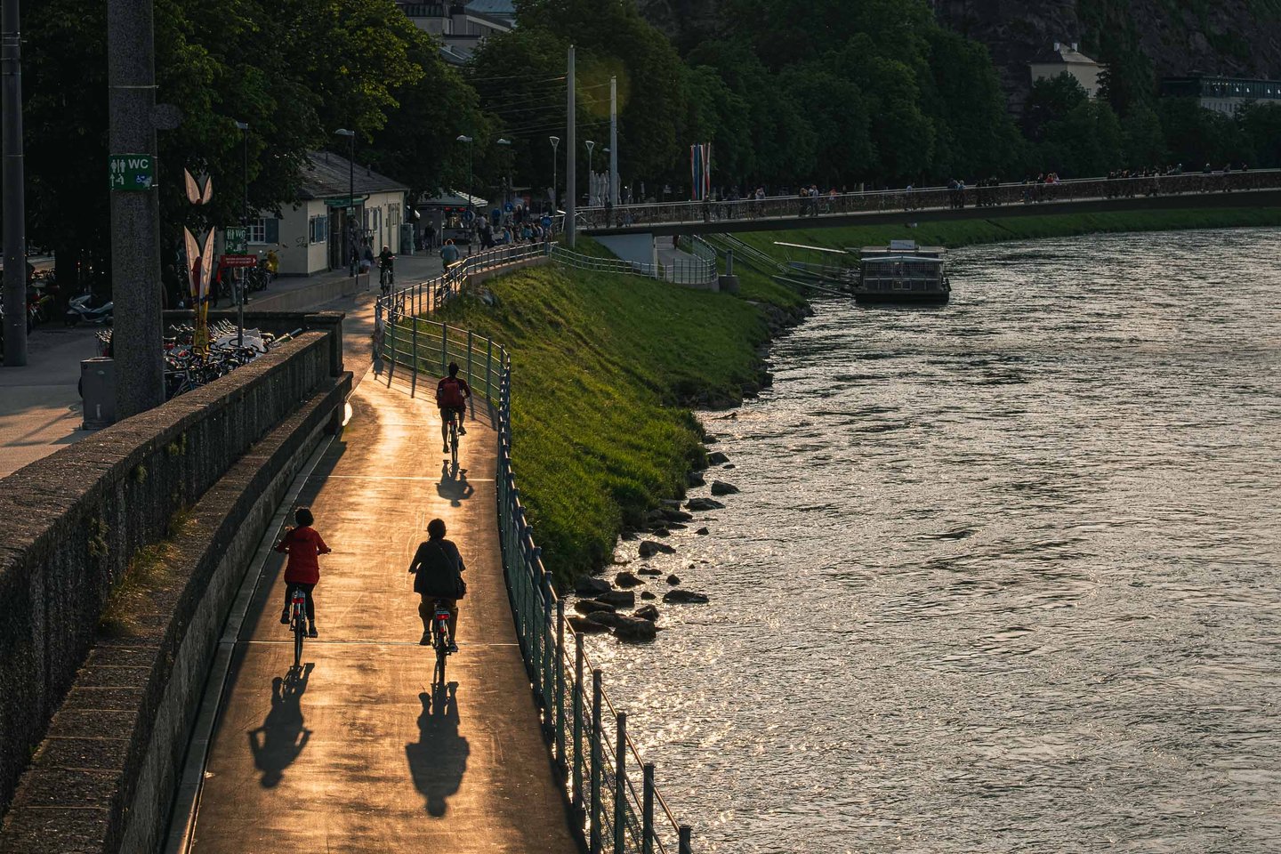 Cyclists on the Salzach-Promenade riverside cycle path at evening with Hohensalzburg Fortress on the hill above the Altstadt across the river