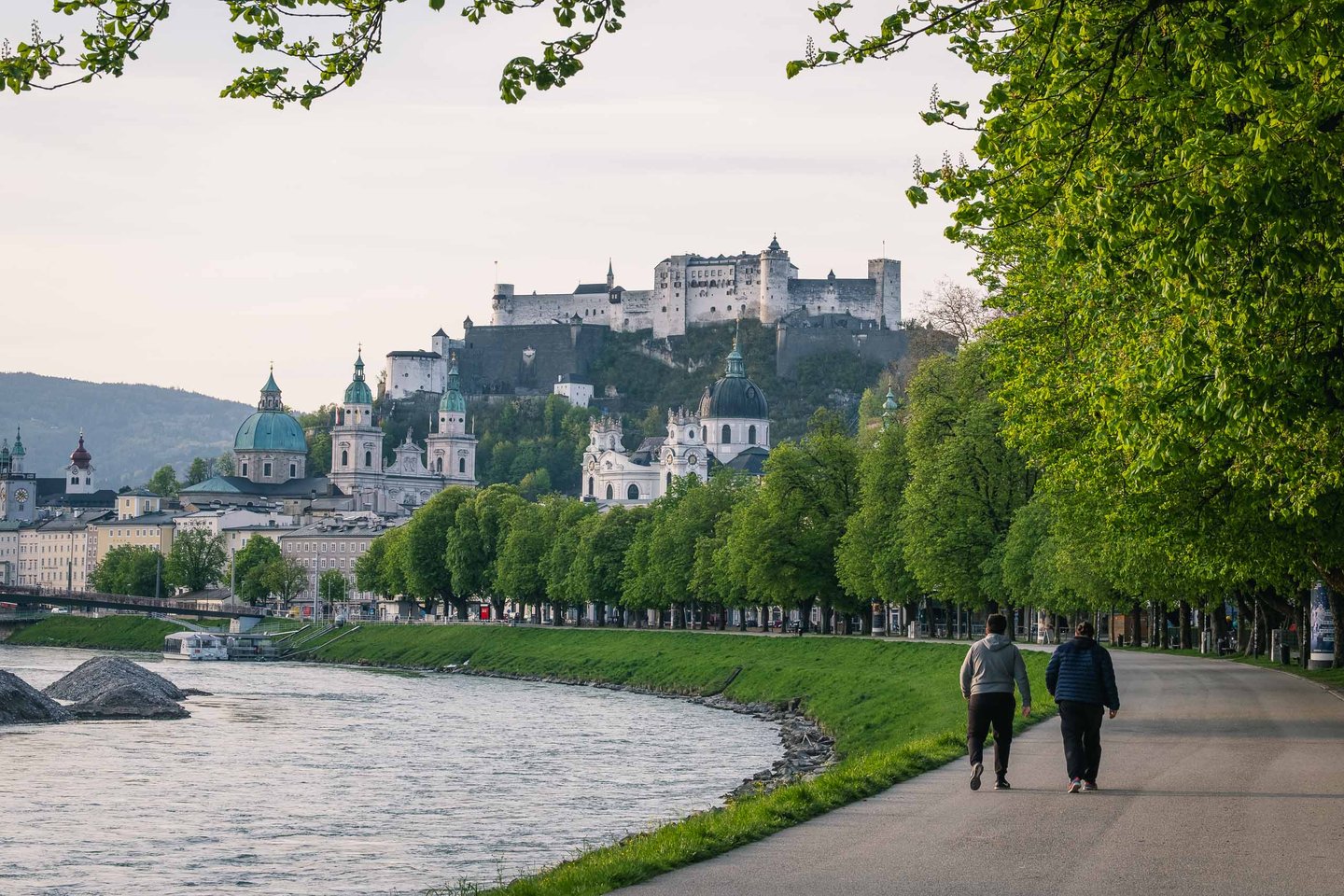 Walkers on the Salzach riverside promenade with Hohensalzburg Fortress rising above Altstadt in Salzburg.