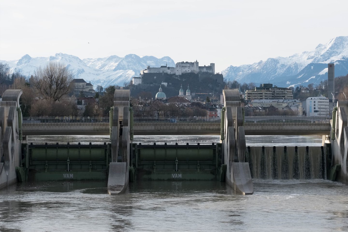 Salzach river weir with Hohensalzburg Fortress and cathedral domes framed by snowy mountains in Salzburg.